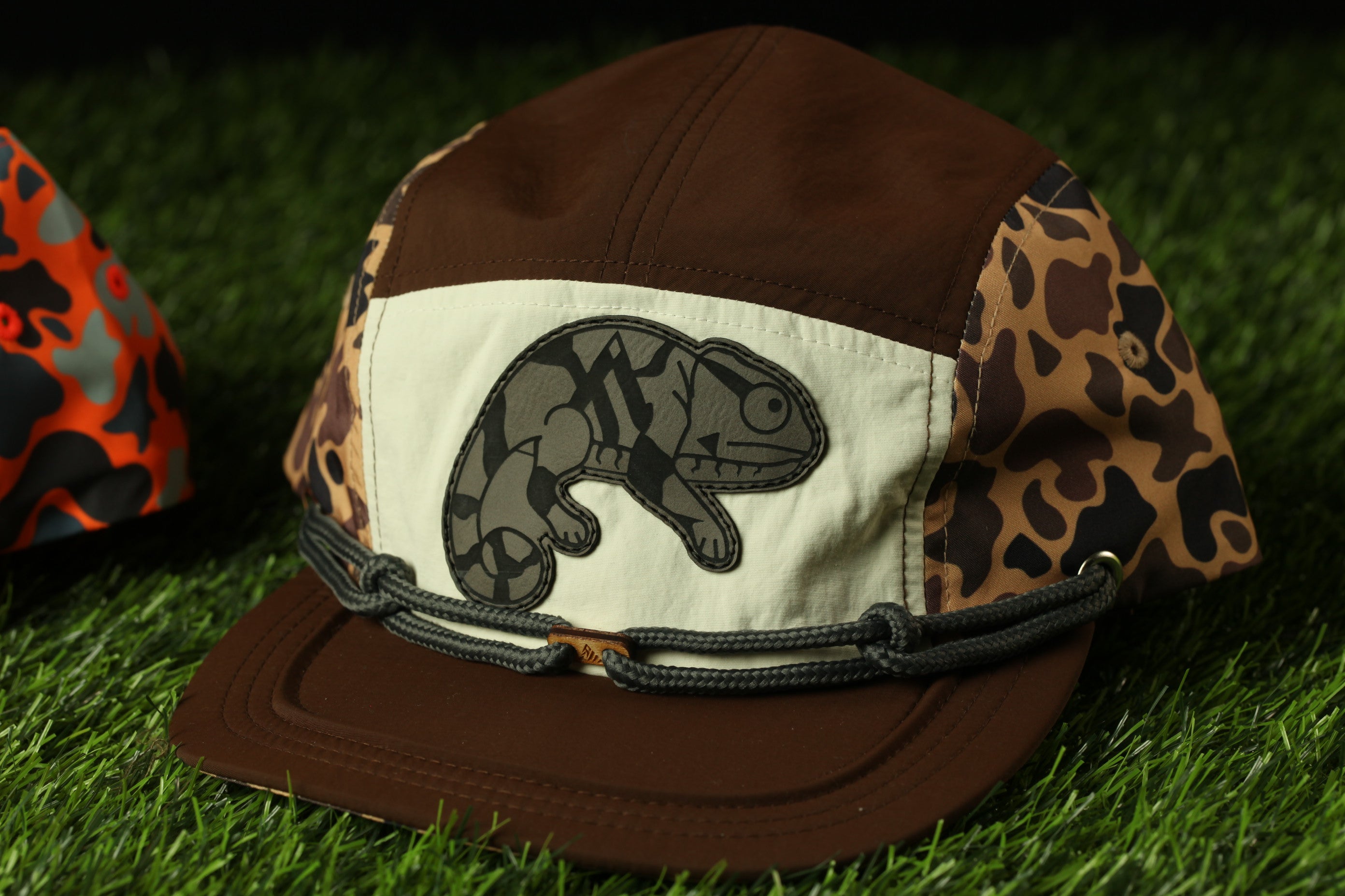 A brown and white baseball cap with a leopard print pattern and a chameleon design on the front, placed on a grassy surface.