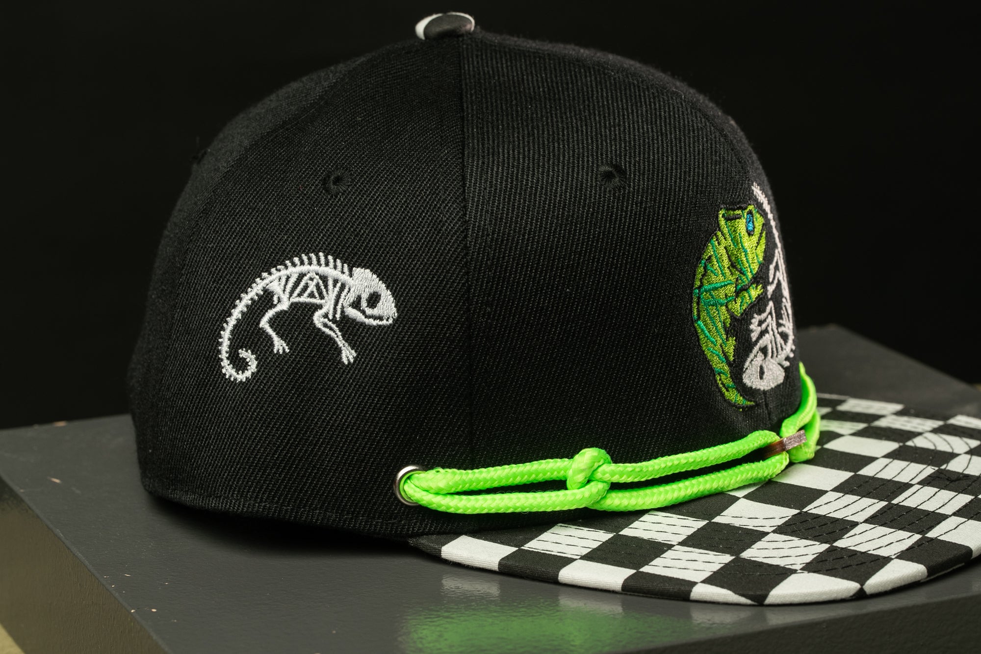 A black baseball cap with a checkered pattern and a white chameleon embroidered on it, along with a green string or strap.