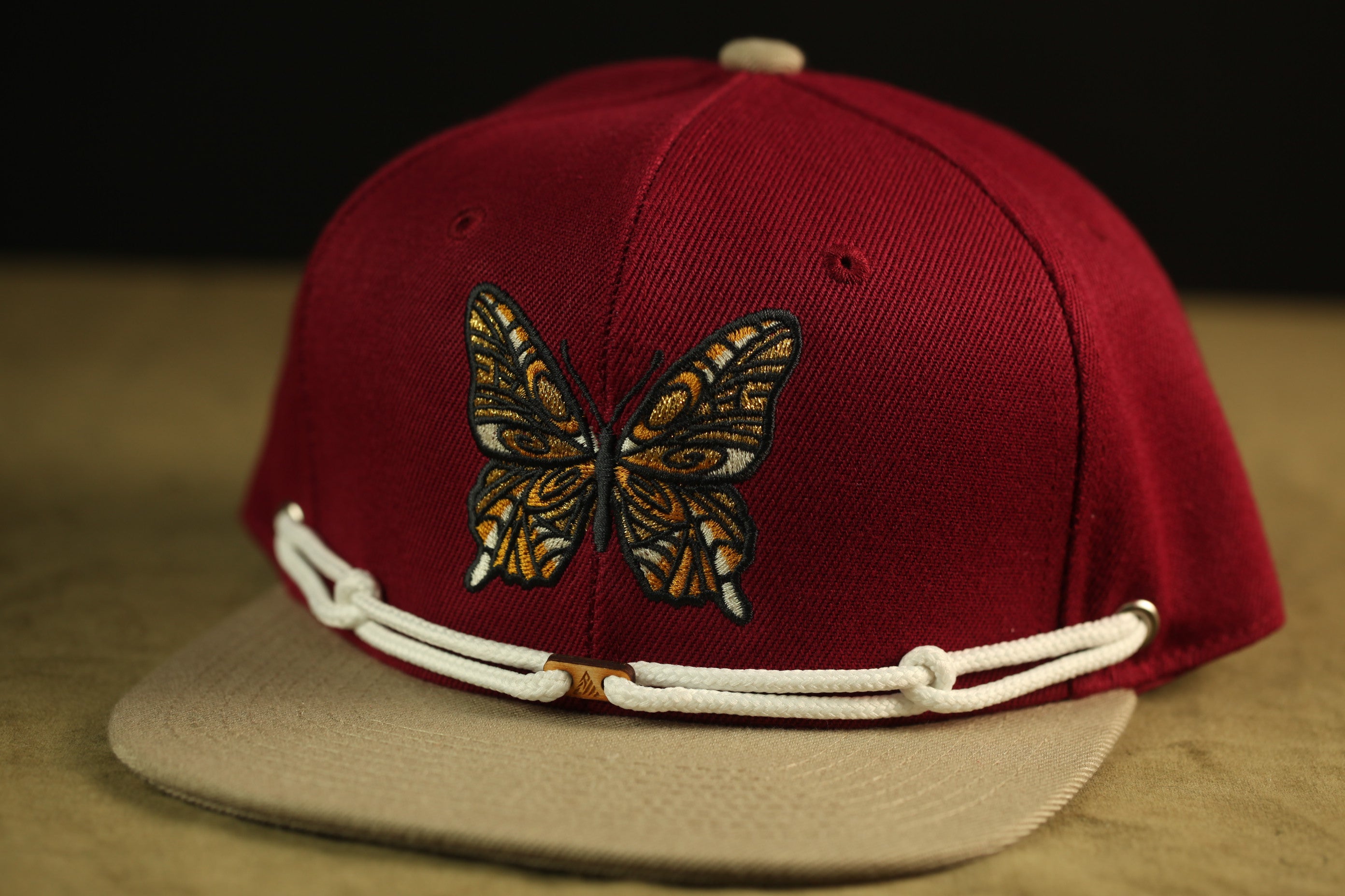 A maroon baseball cap with a white rope trim and a detailed butterfly embroidered on the front.