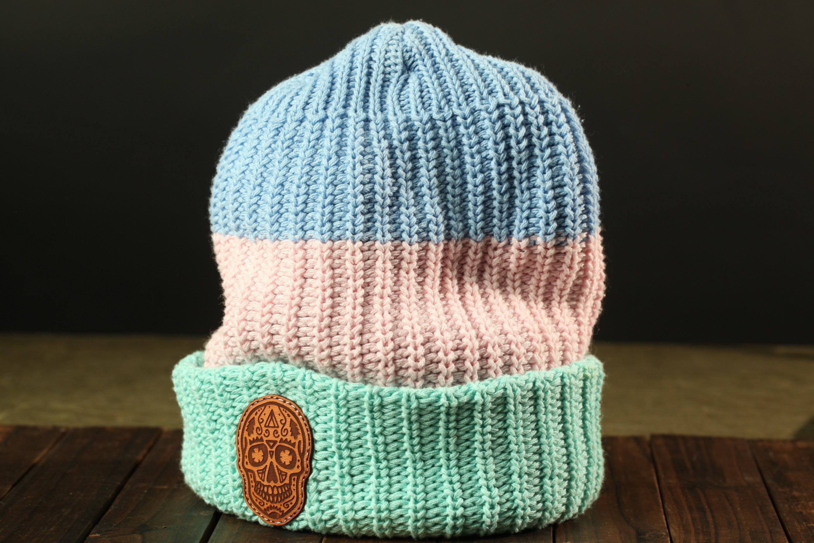 A knitted beanie with a blue and pink striped pattern, featuring a small embroidered skull patch on the front.