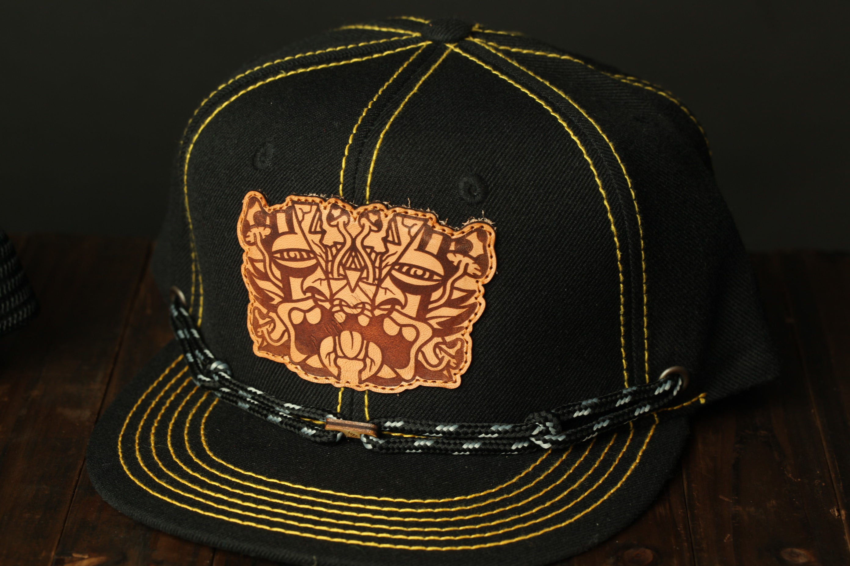 A black baseball cap with a yellow stitching pattern and a brown leather patch featuring a stylized design on the front.