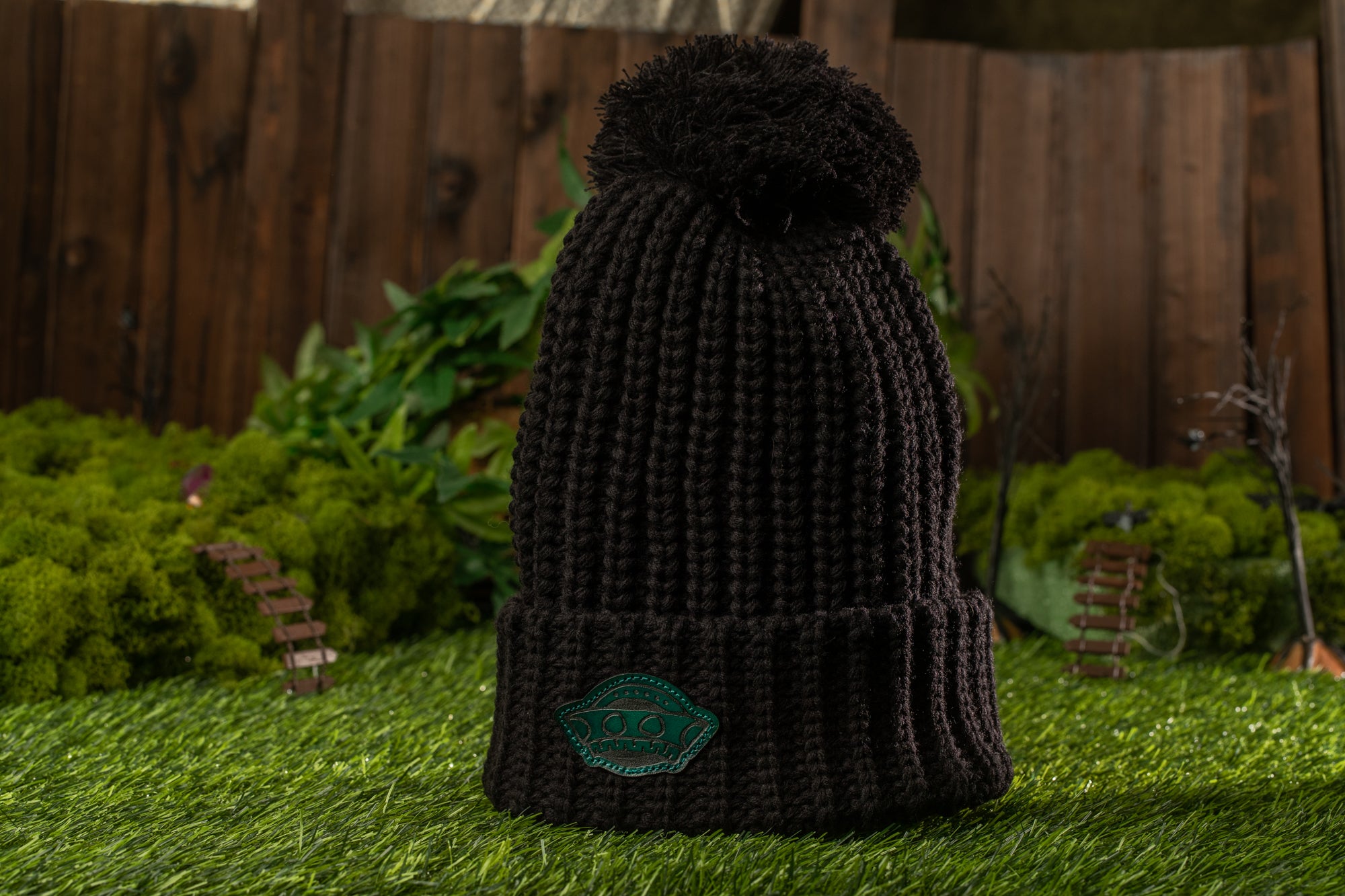 A black knit beanie with a green patch on the front is placed on a grassy surface, with a wooden fence and miniature figurines in the background.