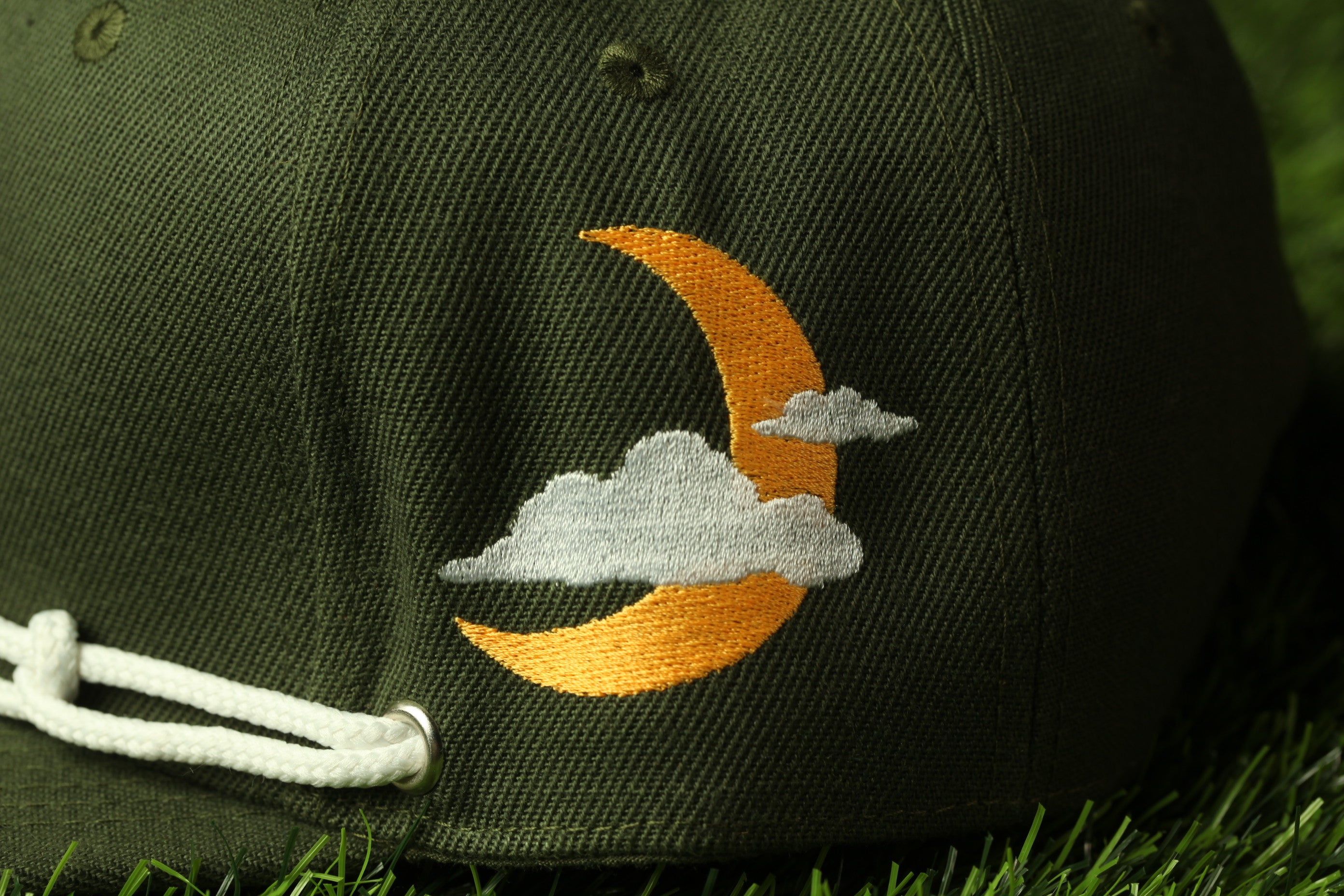 A close-up view of a green baseball cap with an embroidered design depicting a crescent moon and clouds on the front.