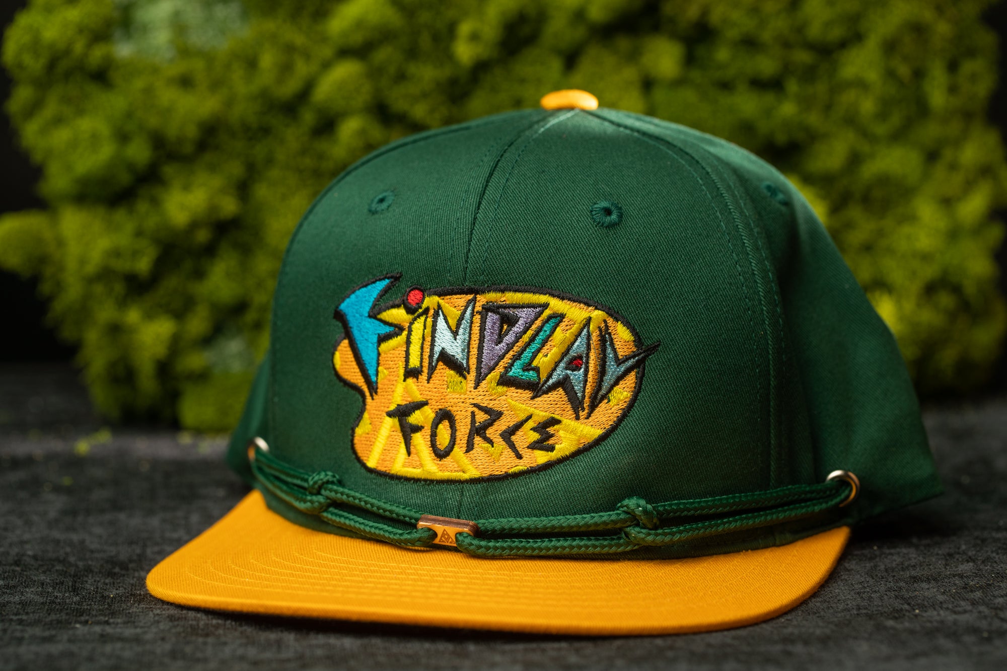 A green baseball cap with a yellow brim and a colorful logo on the front, placed against a background of green moss.