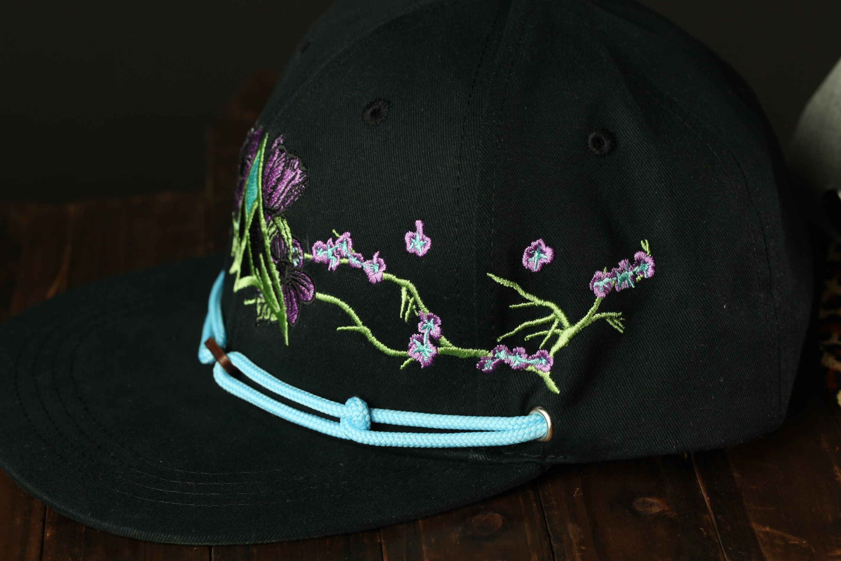 A black baseball cap with a floral embroidered design on the front, featuring purple and green flowers.