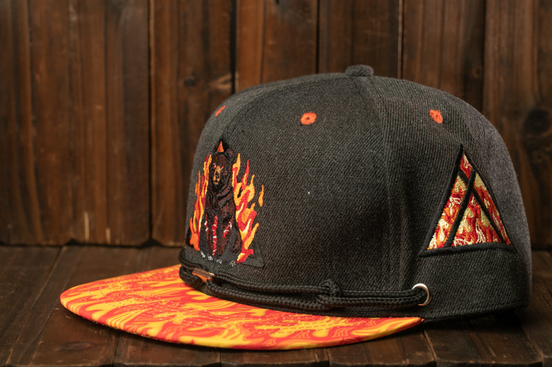 A black baseball cap with a yellow and orange patterned brim, featuring a small embroidered image of a bear and flames on the front.