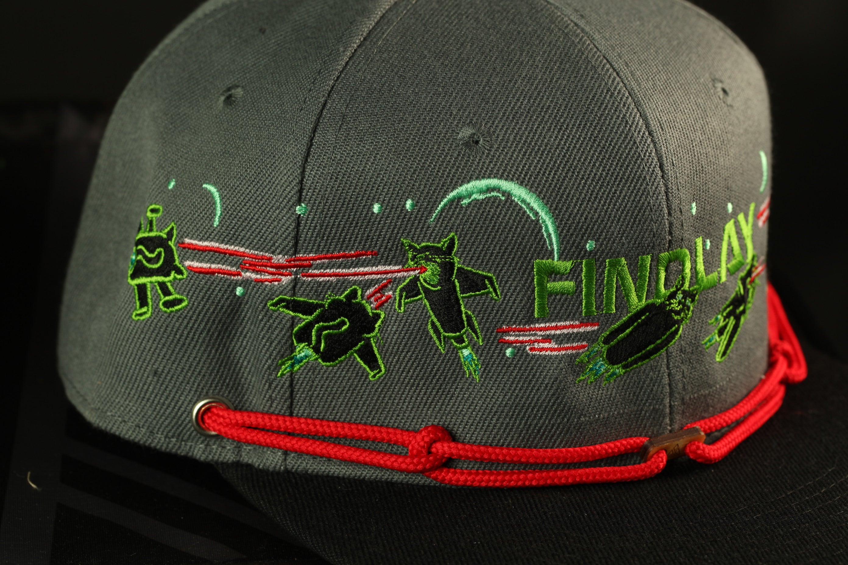 a gray baseball cap with a red string and various green and black designs on it.