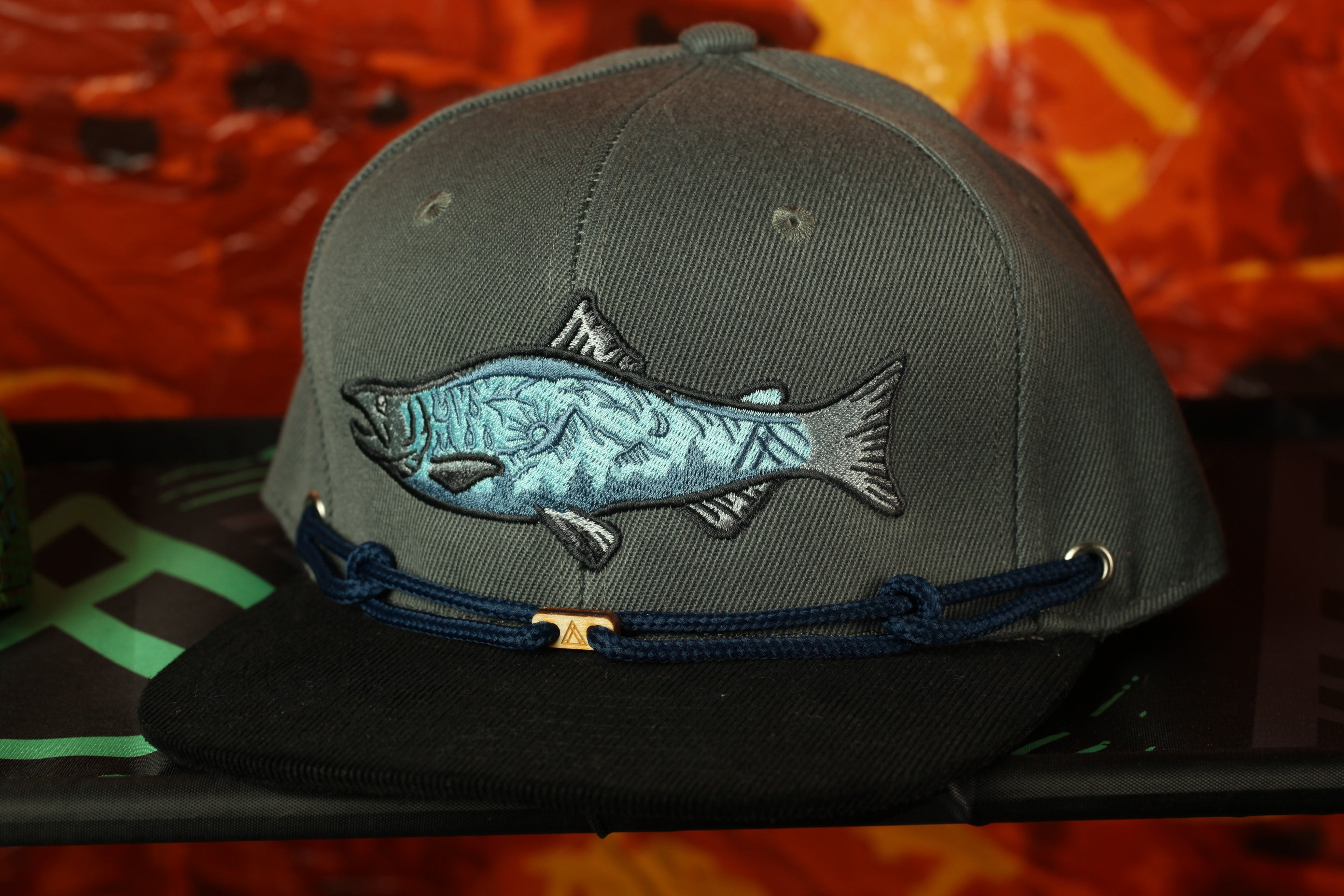 A gray baseball cap with a fish embroidered on it, displayed against a red and orange background.