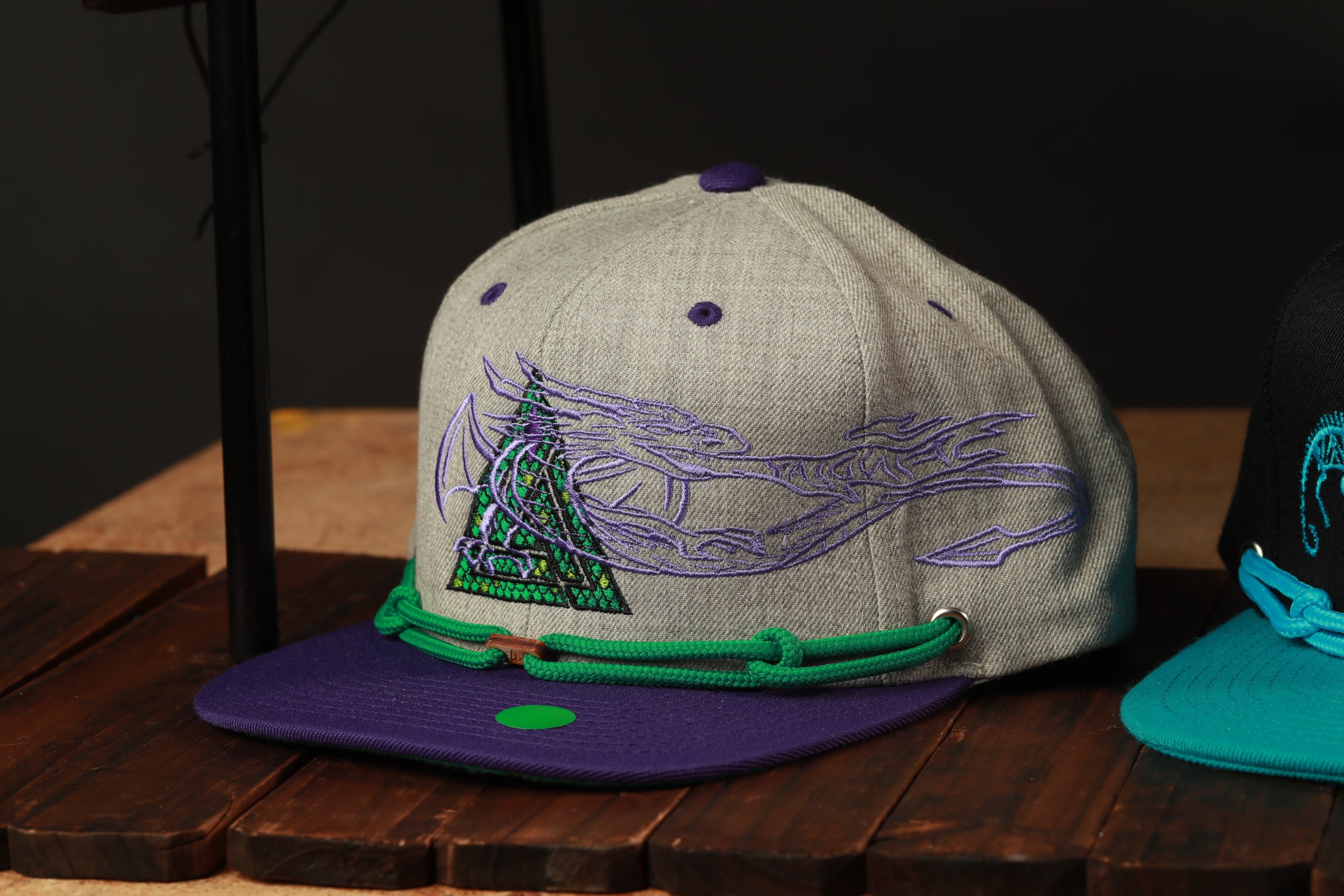 A gray baseball cap with a purple and green design, featuring a dragon-like creature, is displayed on a wooden surface.