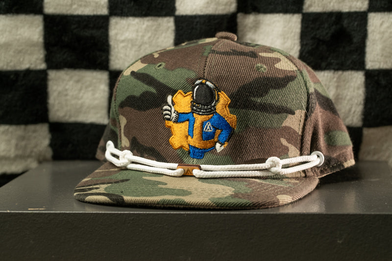 A camouflage baseball cap with a yellow and blue embroidered character on it, sitting on a gray surface against a checkered background.