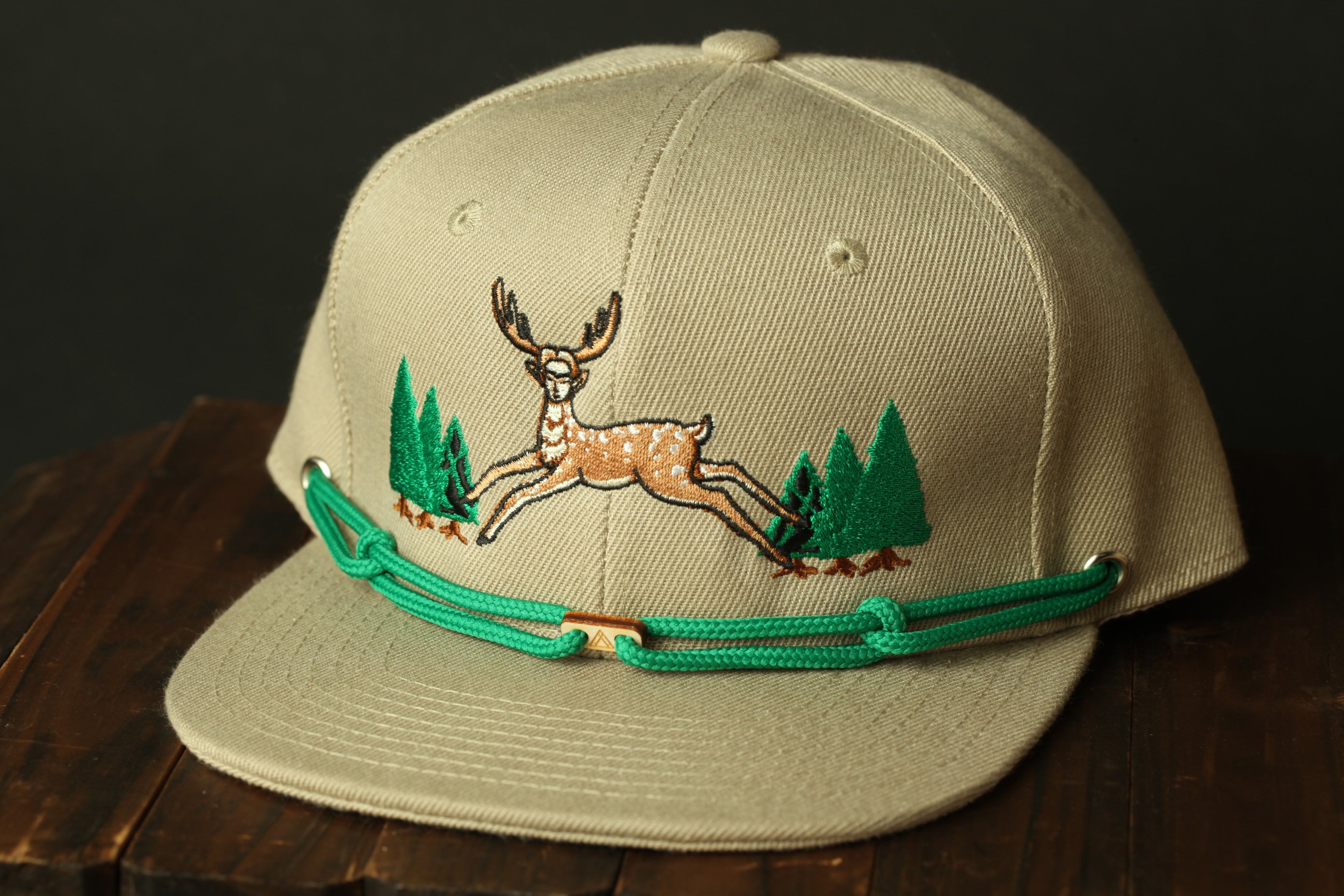 A beige baseball cap with a green embroidered deer and pine trees on the front.