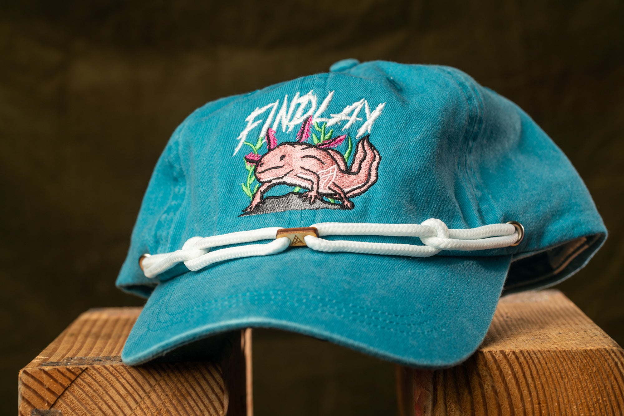 A blue baseball cap with a pink alligator design and the text "FIND LAY" printed on it, sitting on a wooden surface.