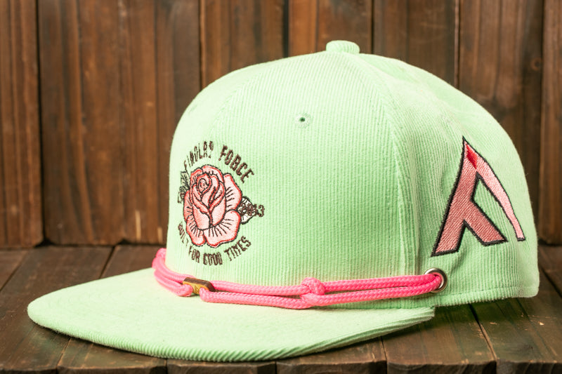A green baseball cap with a pink braided strap and a rose design on the front, placed on a wooden surface.