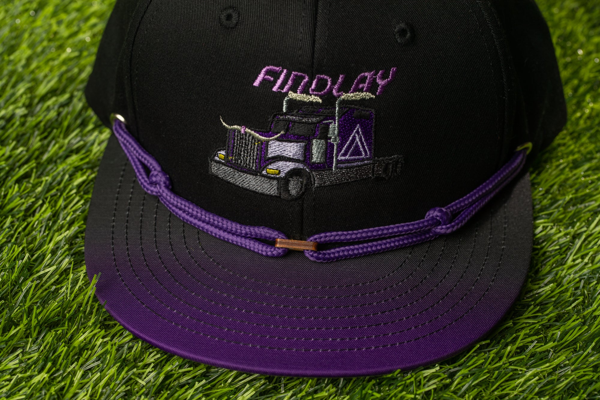 A black baseball cap with a purple strap and an embroidered image of a truck on the front.