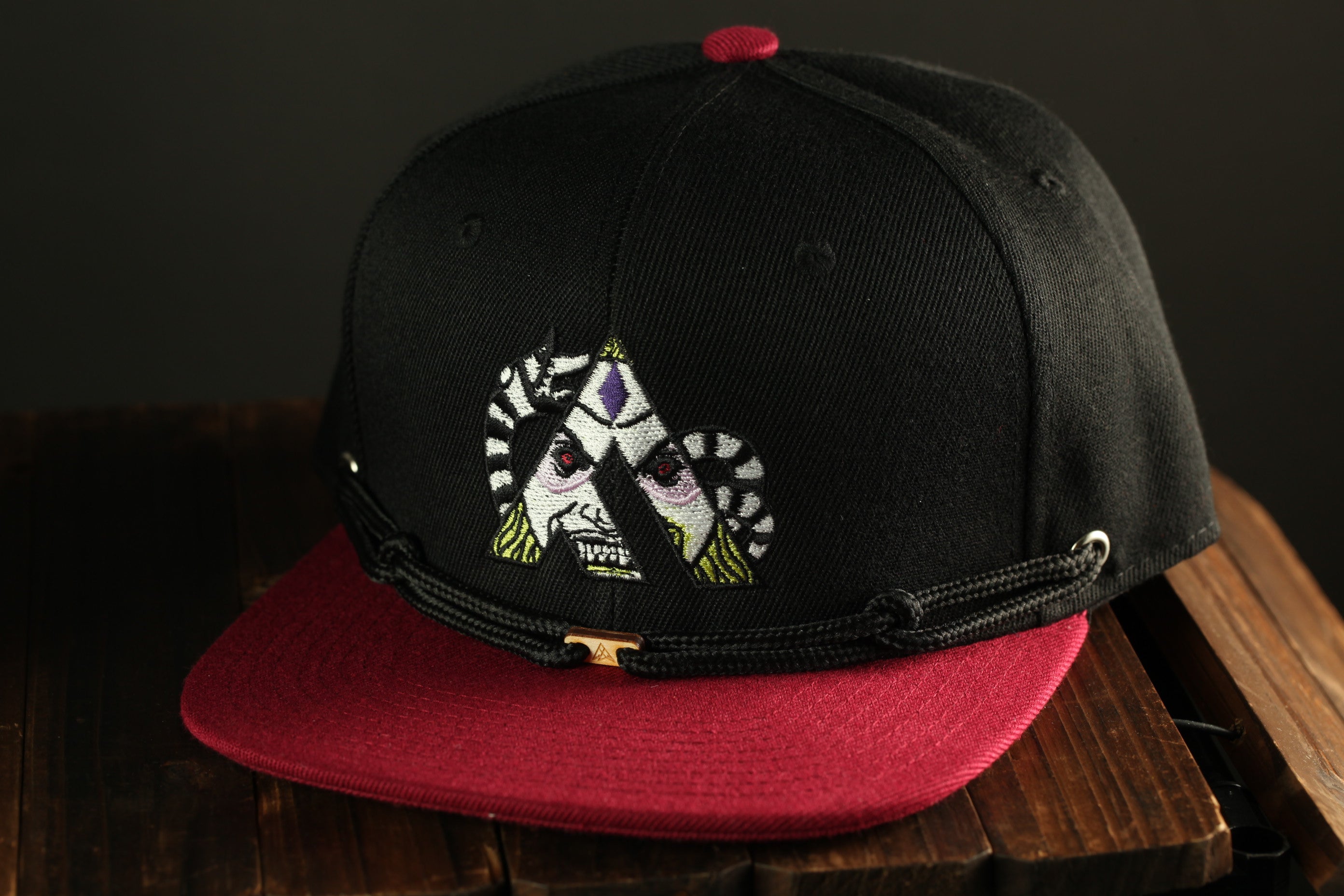 A black baseball cap with a red brim and a graphic design on the front, placed on a wooden surface.