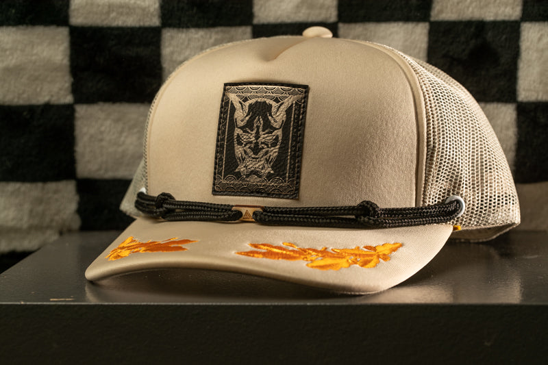 A beige baseball cap with a black and white graphic design on the front, featuring a mask-like figure, is displayed on a black surface against a checkered background.