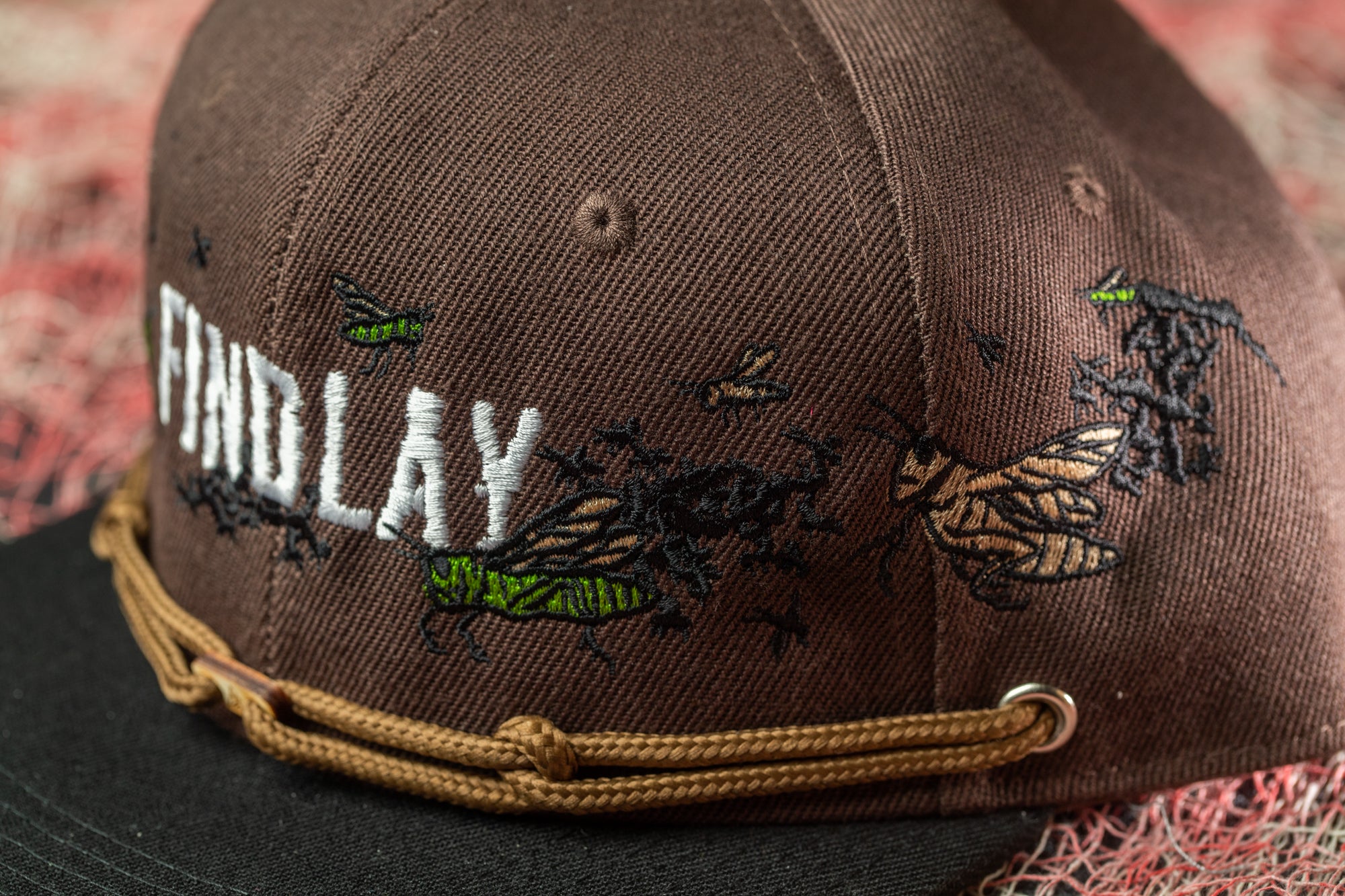 A close-up image of a brown baseball cap with embroidered designs, including a bee and other insects, on the front.