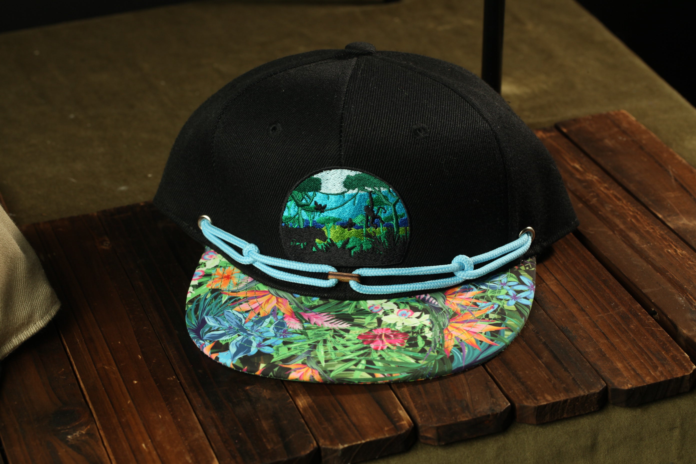A black baseball cap with a colorful floral pattern and a circular patch on the front, placed on a wooden surface.