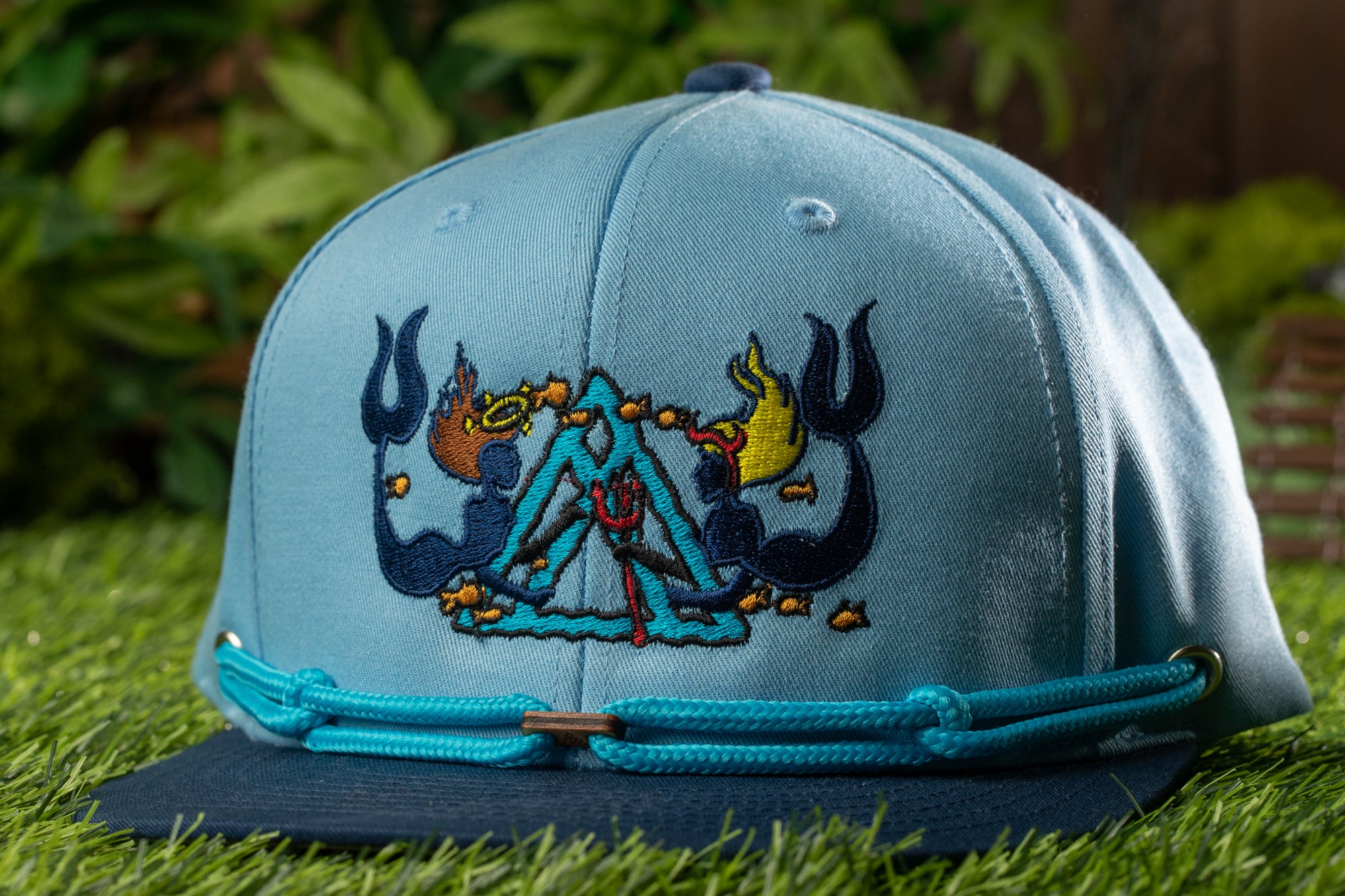 A blue baseball cap with a colorful, embroidered design on the front, sitting on a grassy surface.