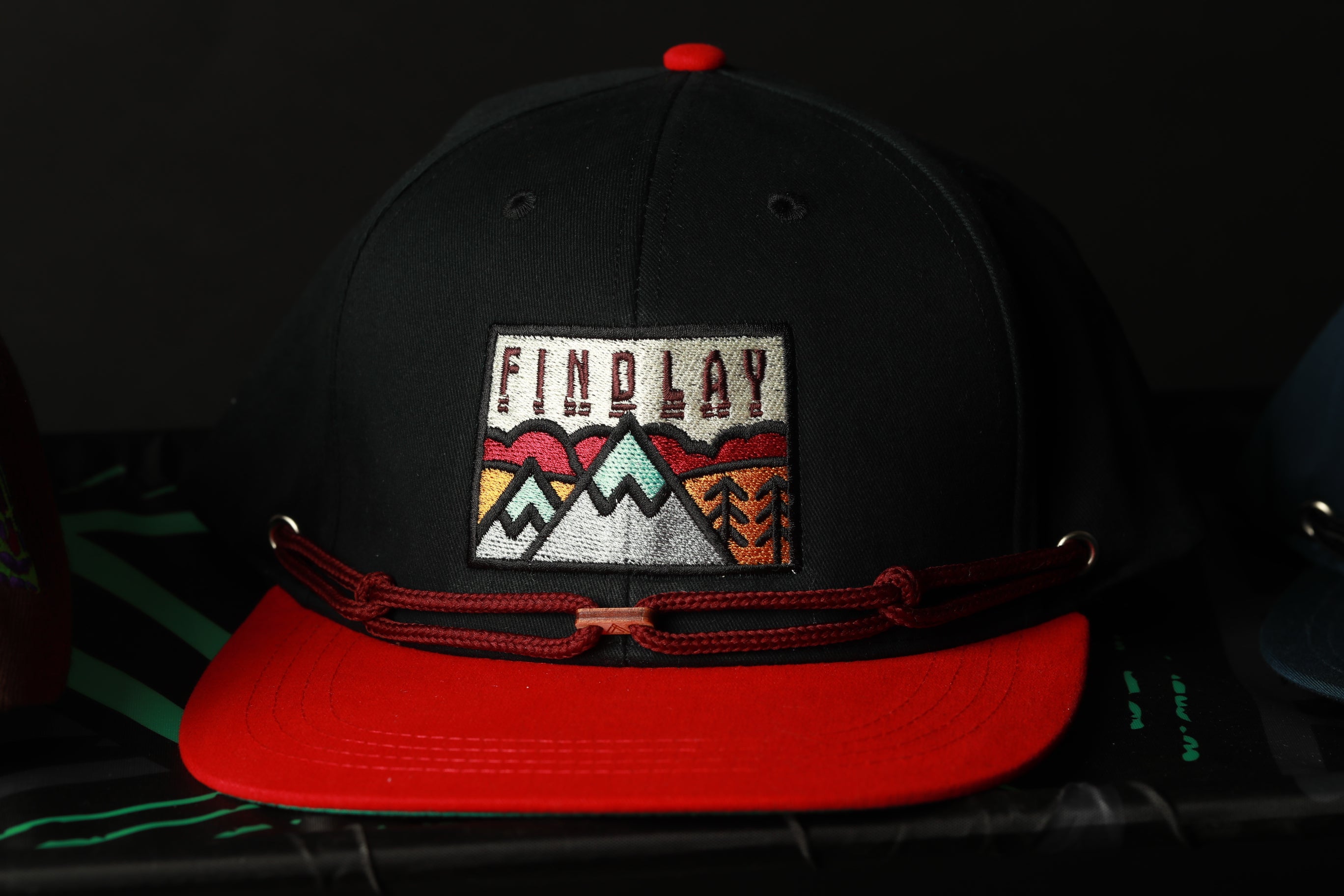 A black baseball cap with a red brim and a patch on the front displaying a mountain landscape.