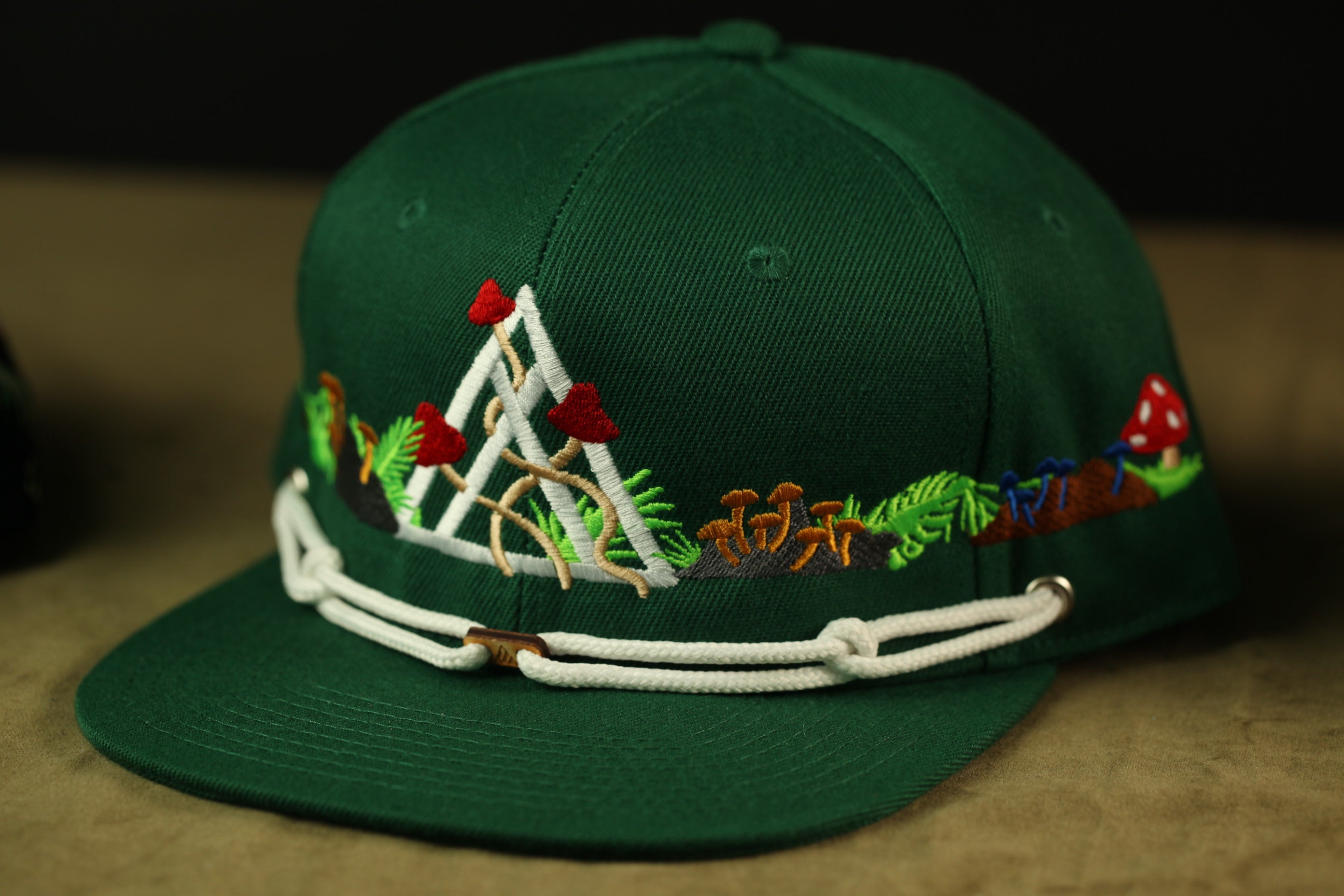 A green baseball cap with a white band and various colorful embroidered designs, including mushrooms, leaves, and a mountain-like shape.