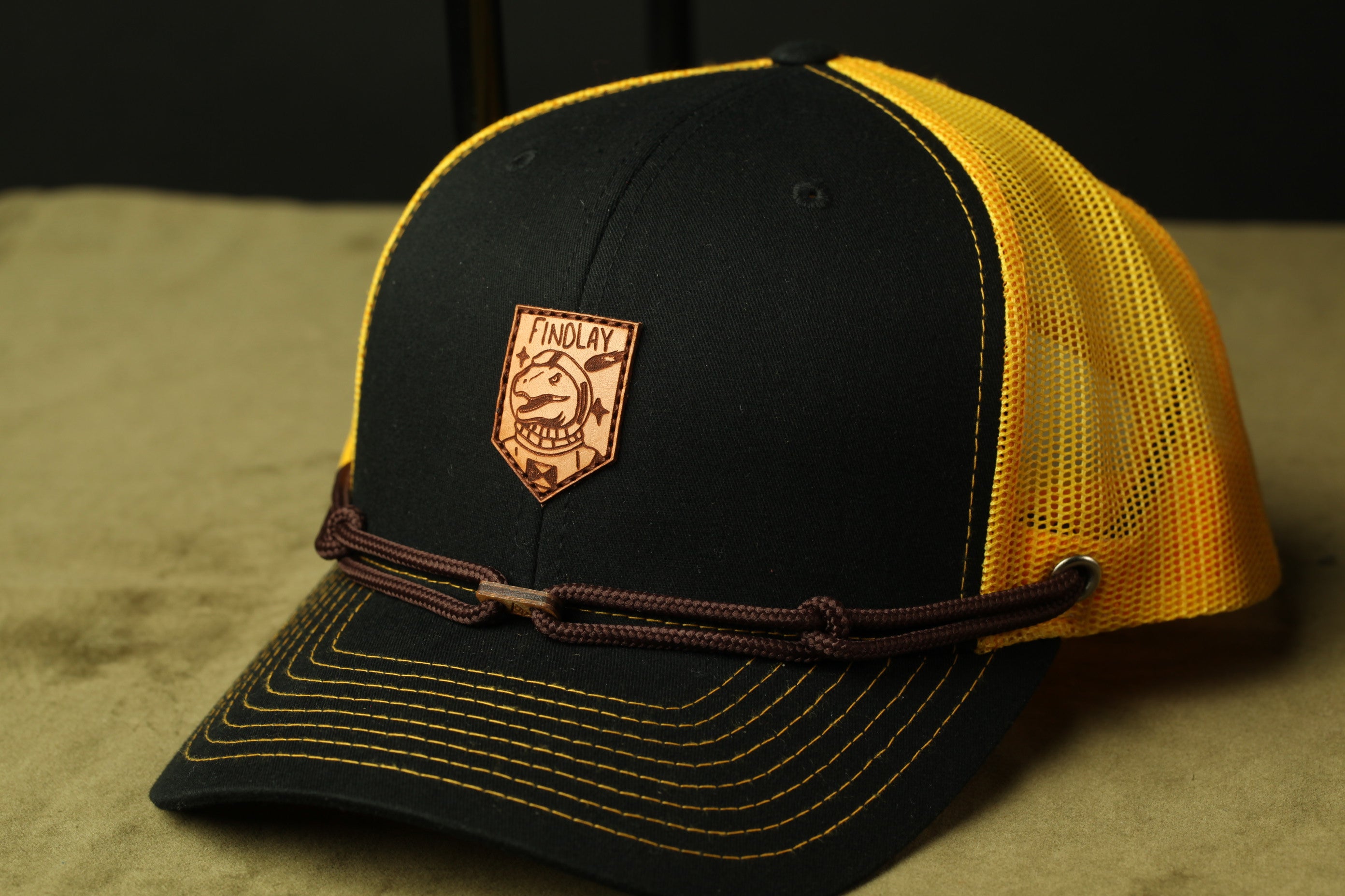 A black baseball cap with a yellow mesh panel, featuring a brown leather patch with a logo on the front.