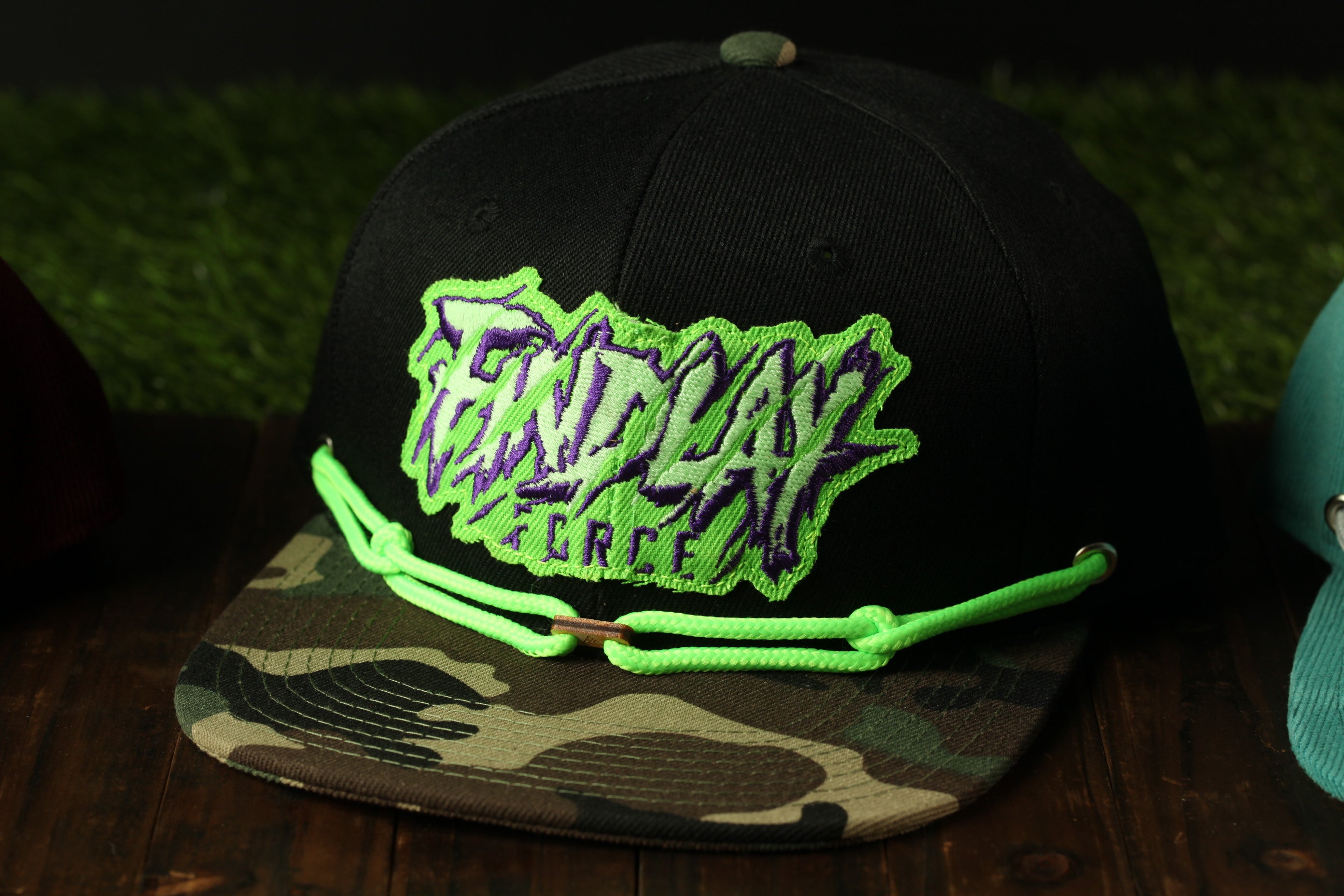 A black baseball cap with a camouflage pattern and neon green strings, featuring a large text logo on the front.