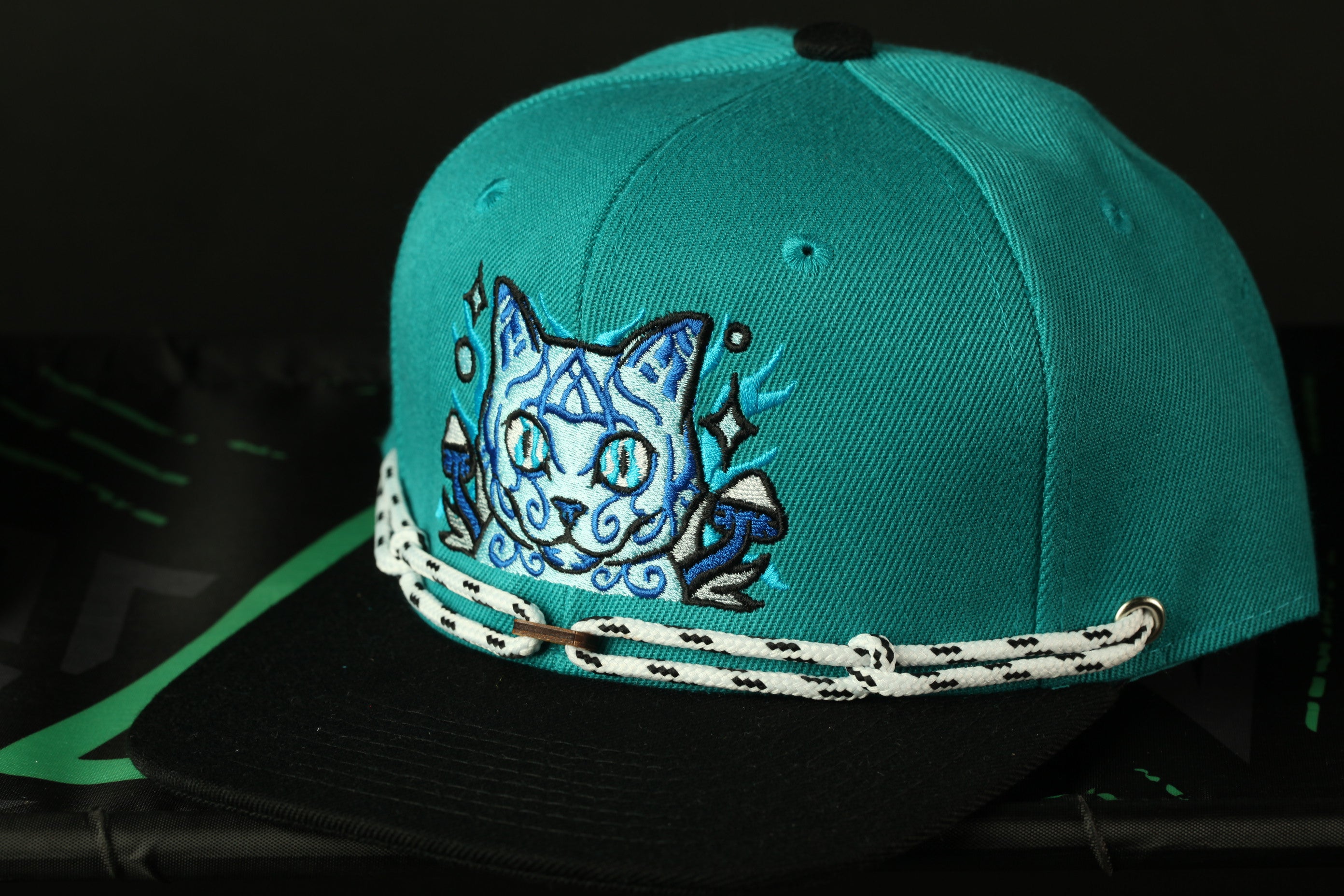 A turquoise baseball cap with a cat embroidered on it, featuring a black brim and white string.
