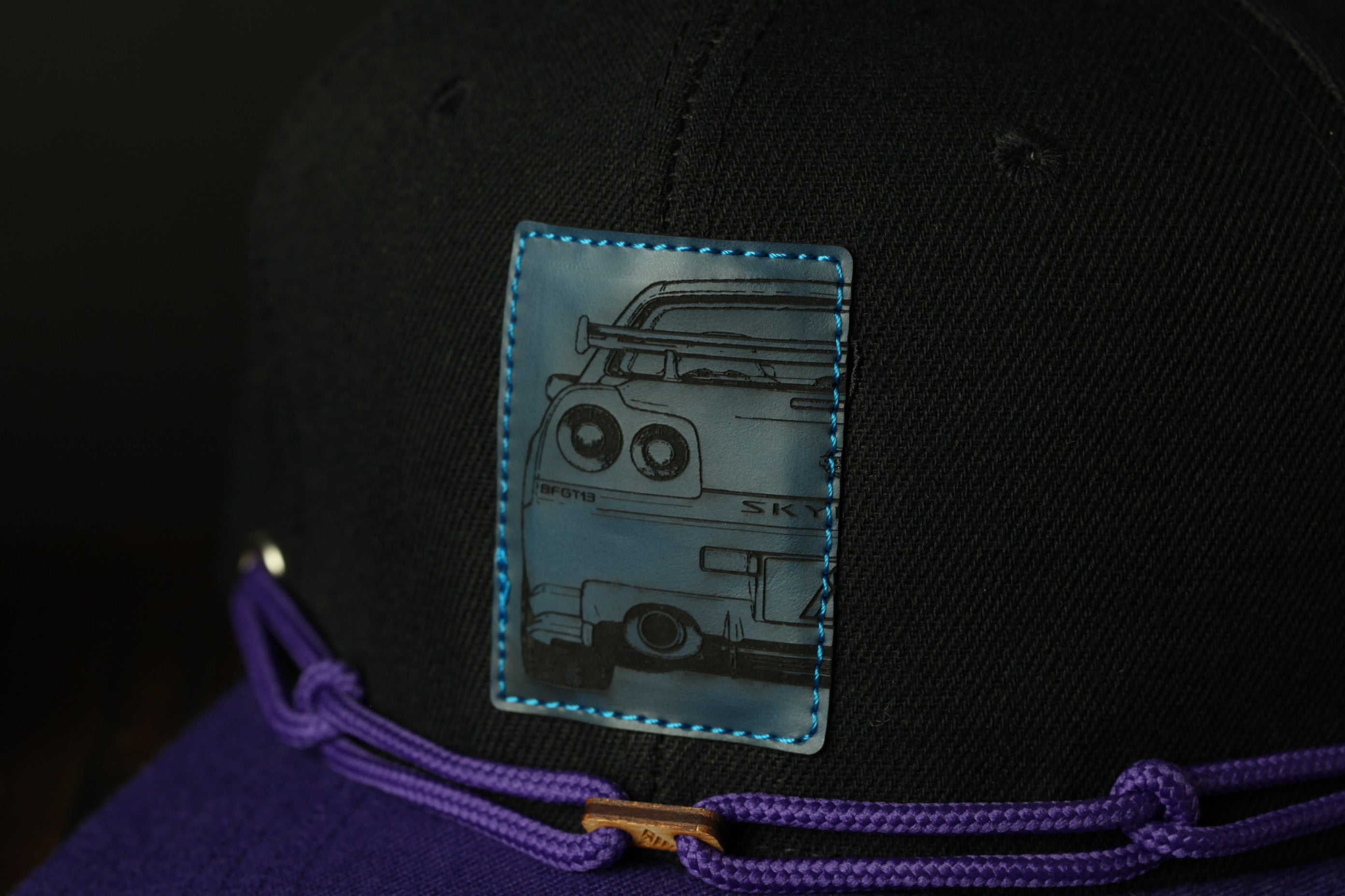 A close-up view of a black baseball cap with a blue patch featuring a graphic of a car.