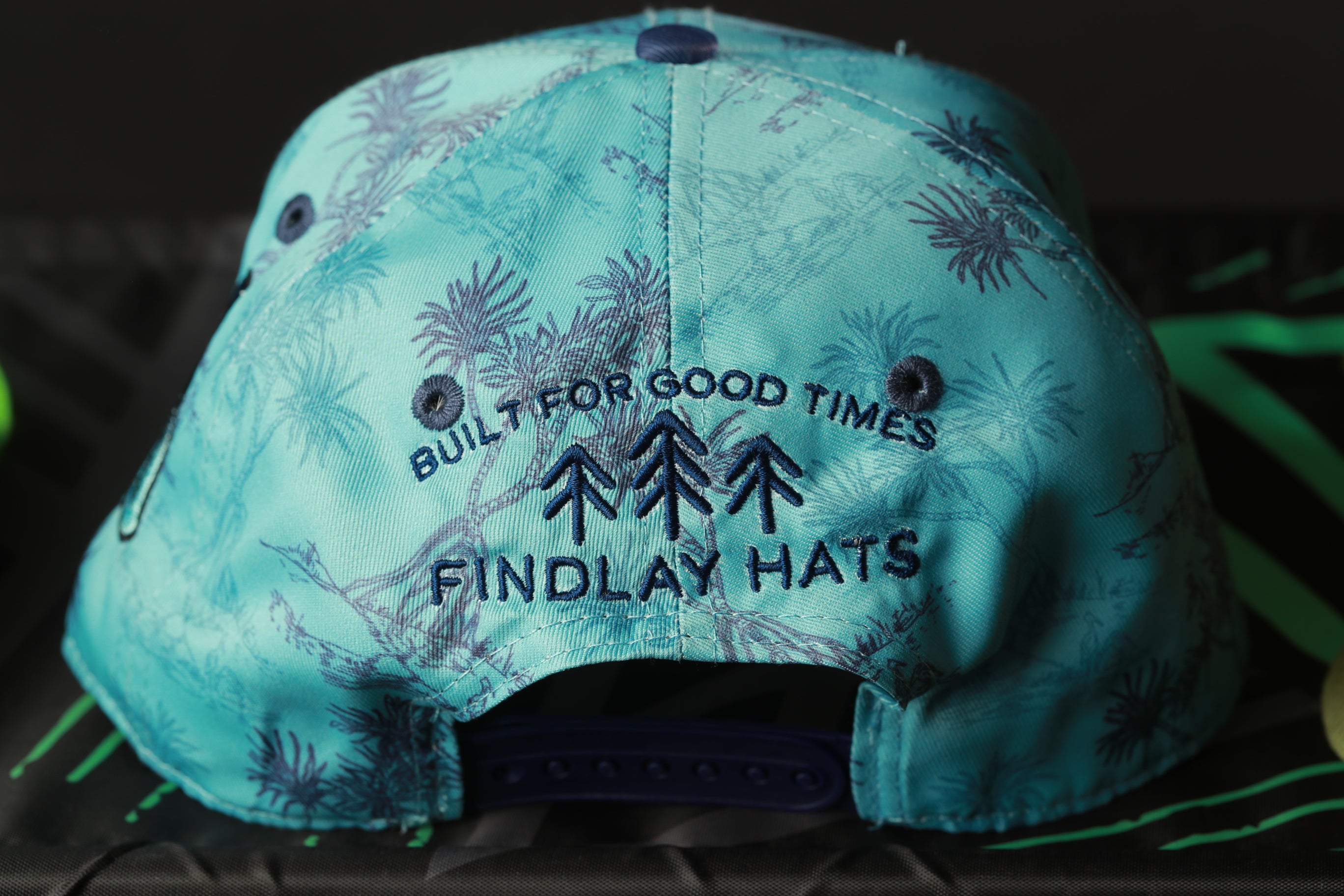 a turquoise baseball cap with a pattern of palm trees and the text "BUILT FOR GOOD TIMES FINDLAY HATS" printed on it.