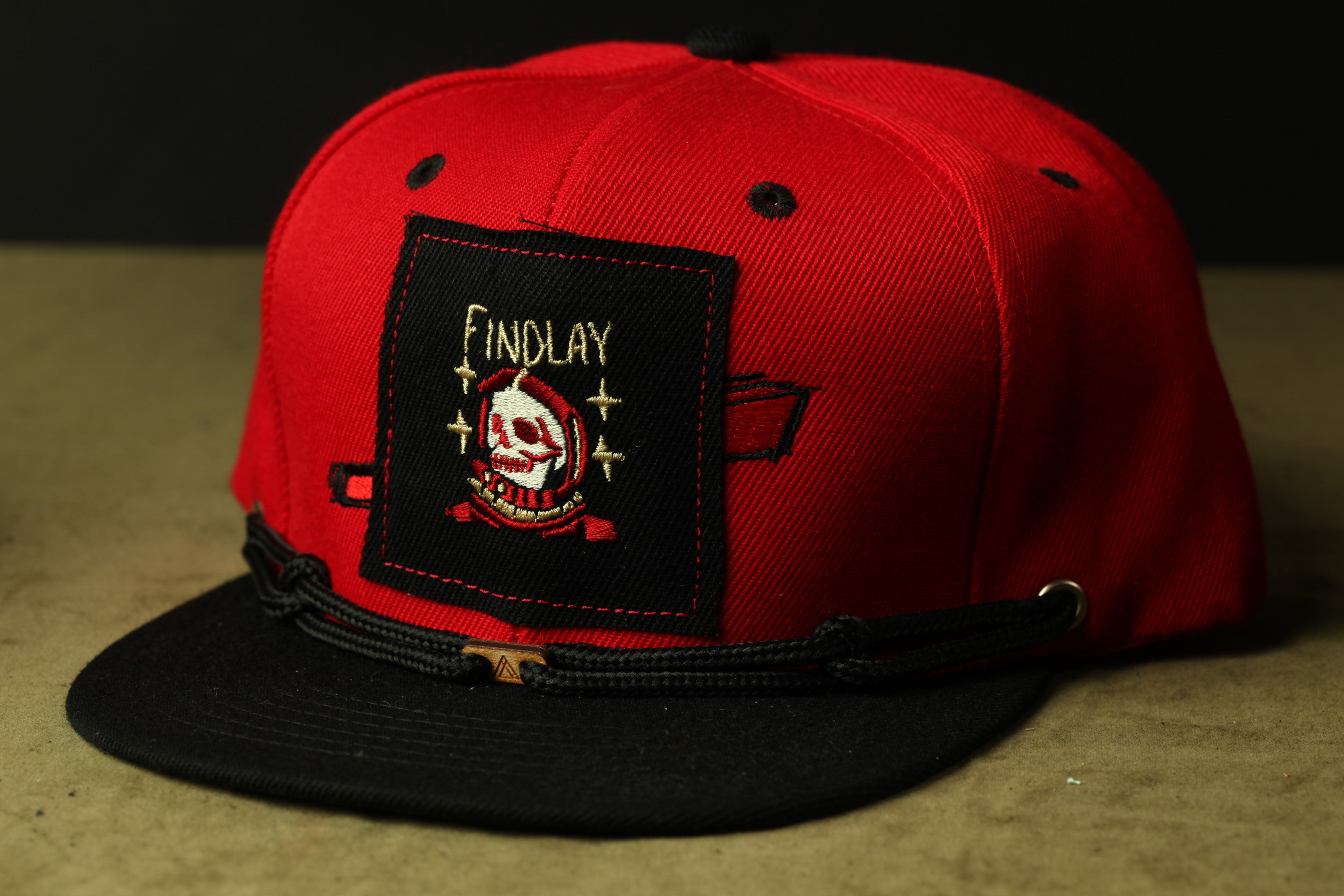 A red baseball cap with a black brim and a black band around the front. The cap has a patch on the front with the text "Findlay" and an image of a skull and crossbones.