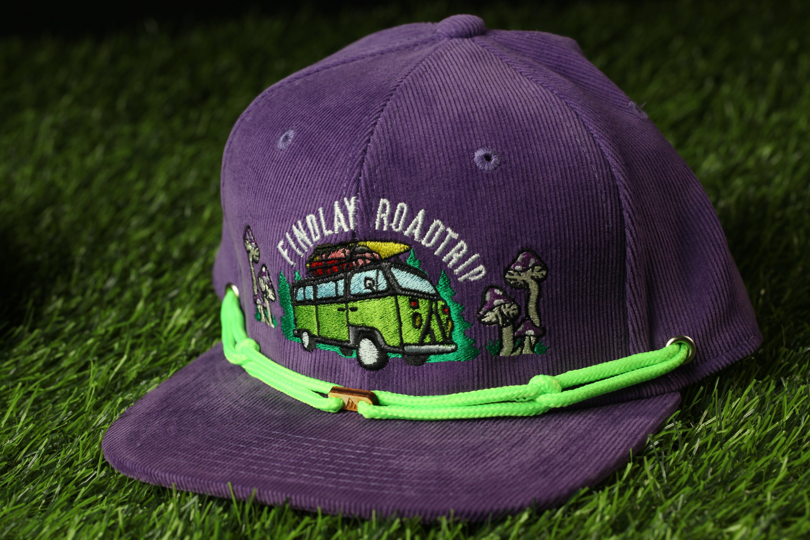 A purple baseball cap with a green string and an embroidered design on it, featuring a van and mushrooms, is placed on a grassy surface.