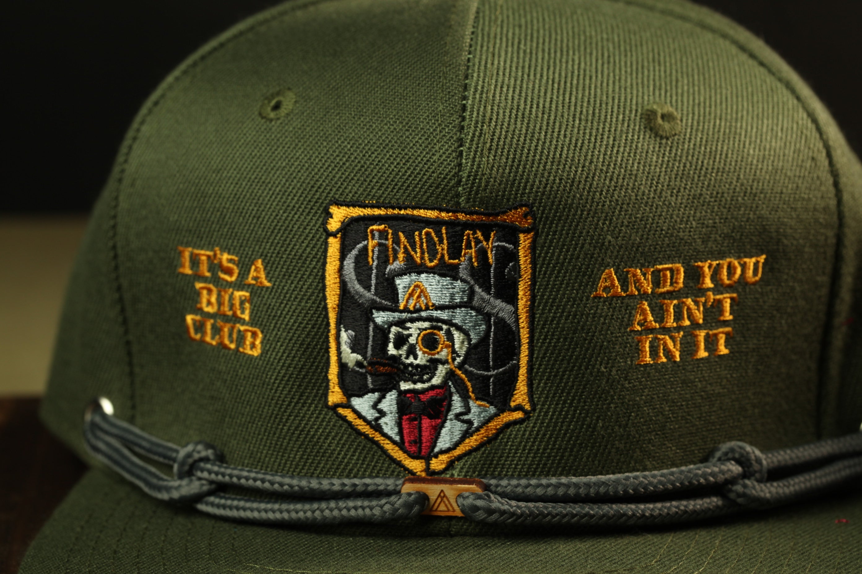 A close-up of a green baseball cap with an embroidered patch featuring a skull wearing a top hat and a cane, along with the text "IT'S A BIG CLUB AND YOU AIN'T IN IT".
