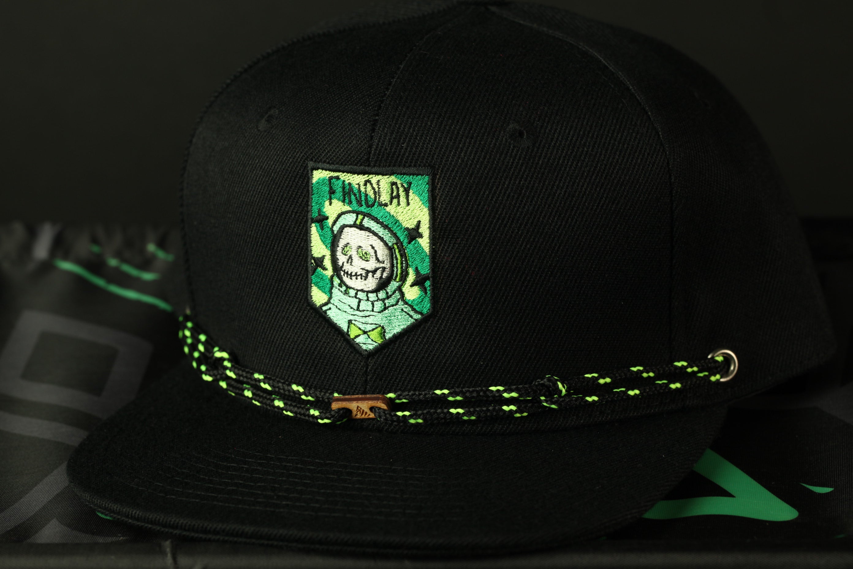 A black baseball cap with a green and yellow patch featuring a skull and crossbones design, along with a green and black braided strap.