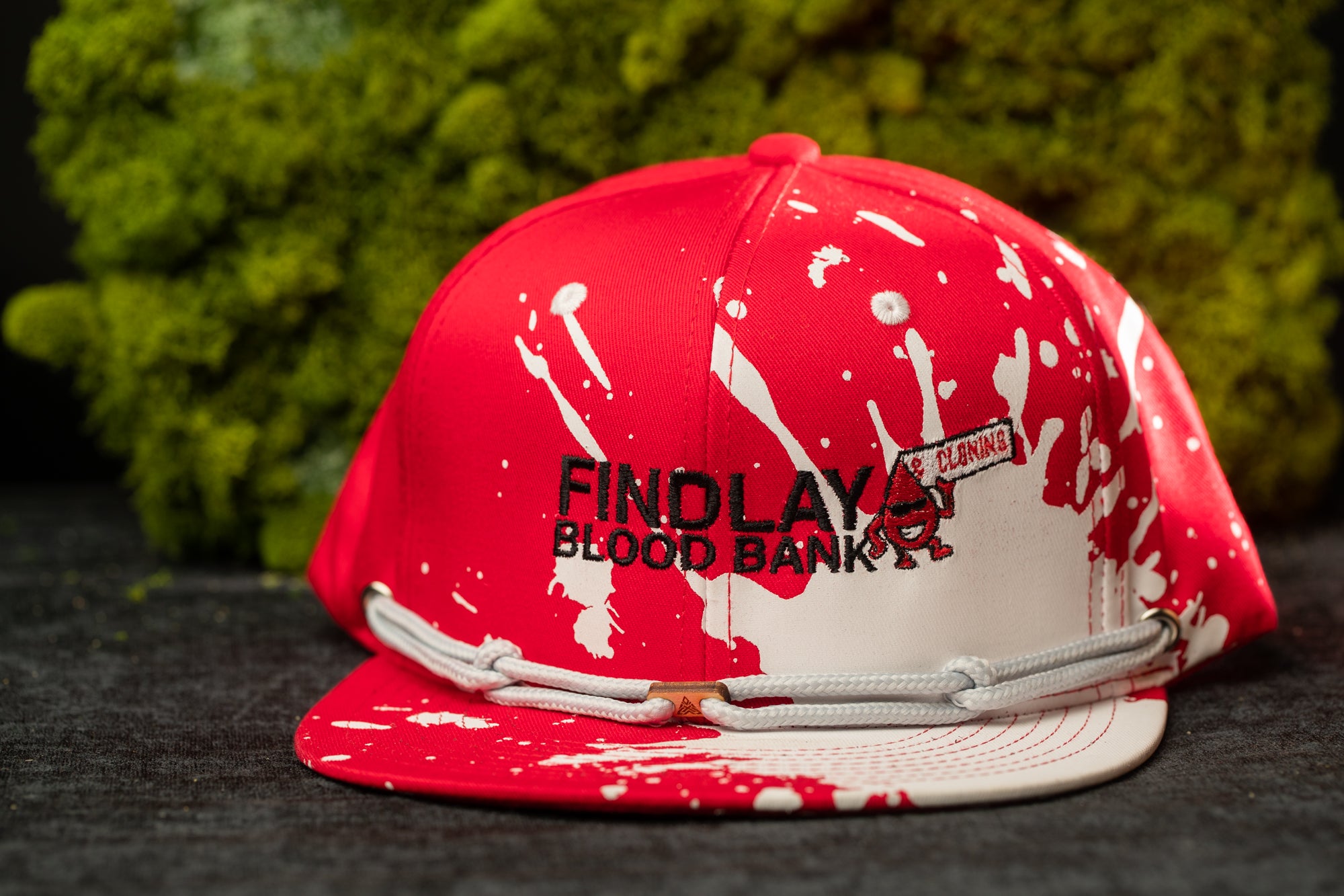 A red baseball cap with white splatters and the text "FIND LAY" printed on it, placed on a black surface against a green background.
