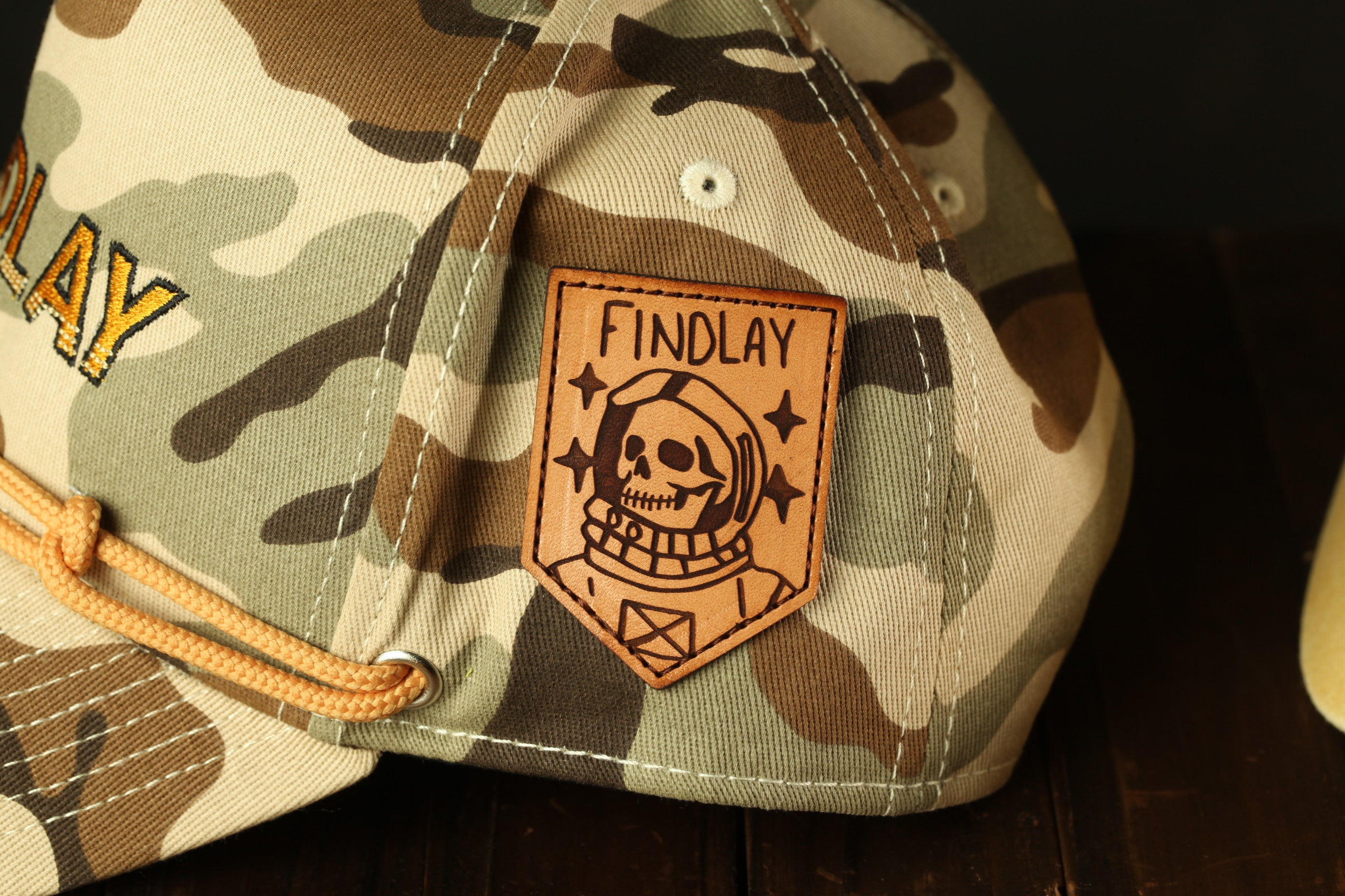 A camouflage baseball cap with a leather patch on the front, featuring a skull and the text "FINDLAY".