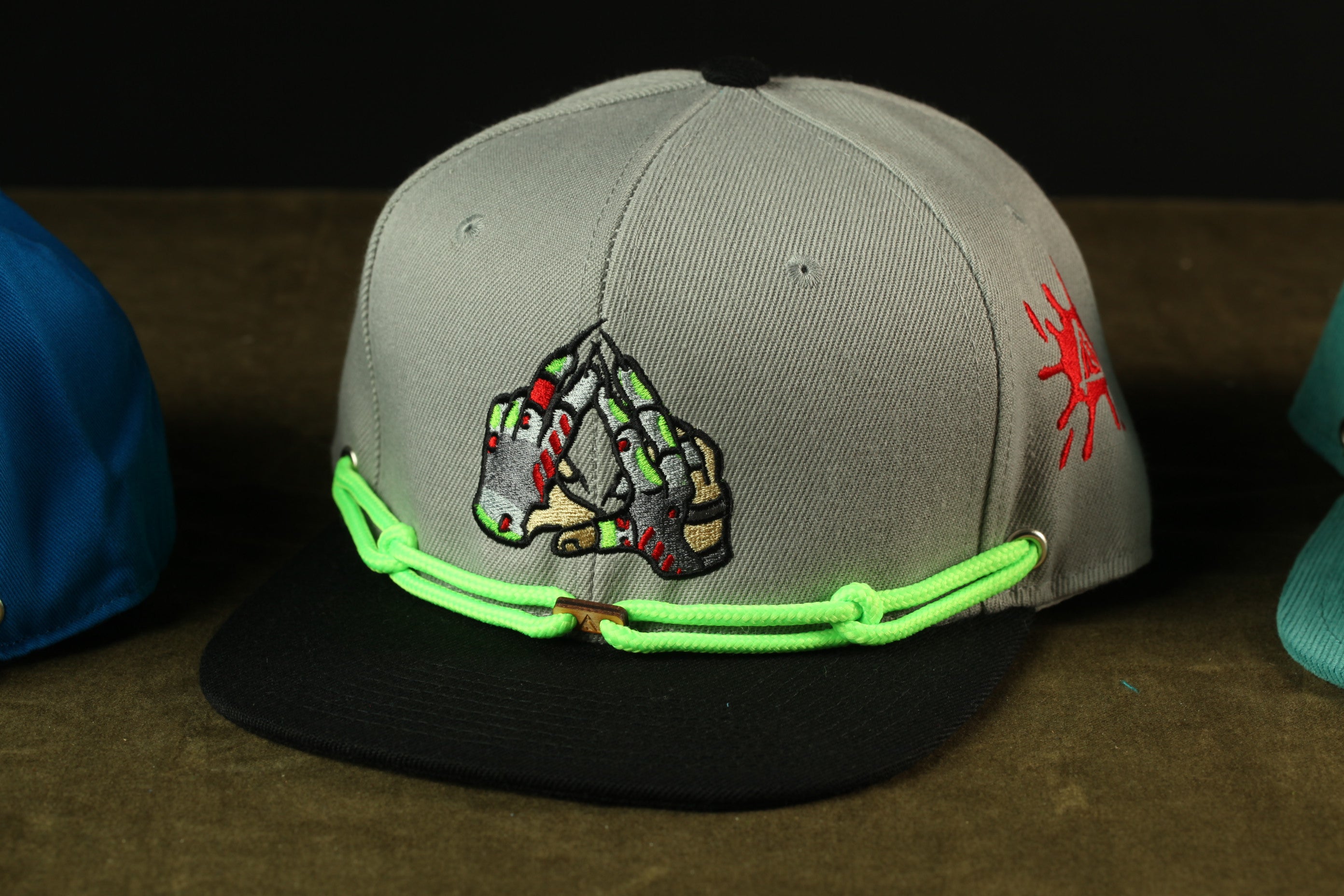 A gray baseball cap with a graphic design on the front, featuring a pair of hands and a skull-like shape. The cap is wearing a green and black strap.