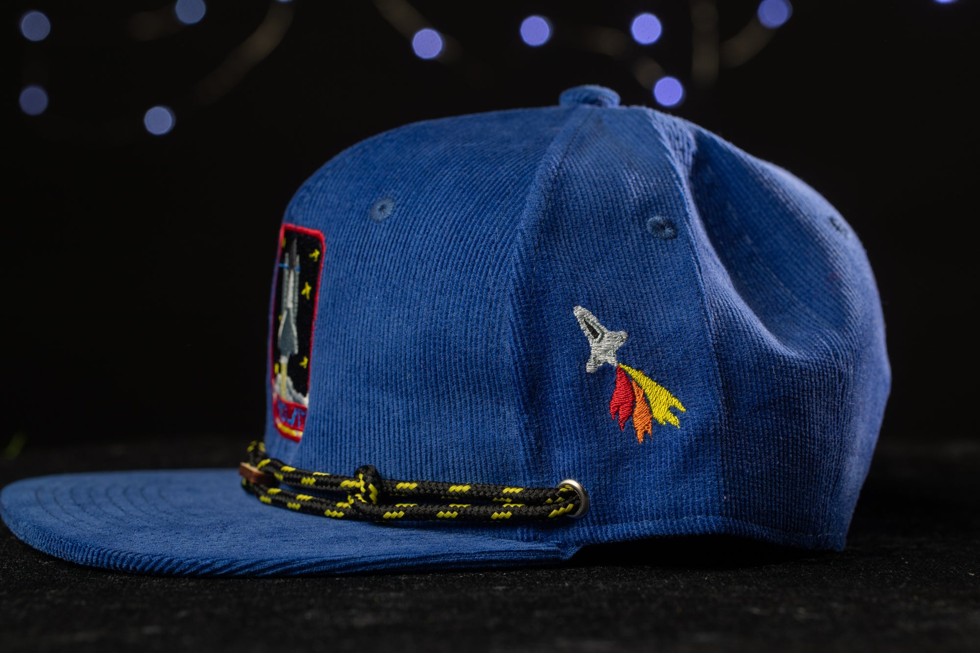A blue baseball cap with a space-themed patch and a rocket design on the front, set against a dark background with twinkling lights.