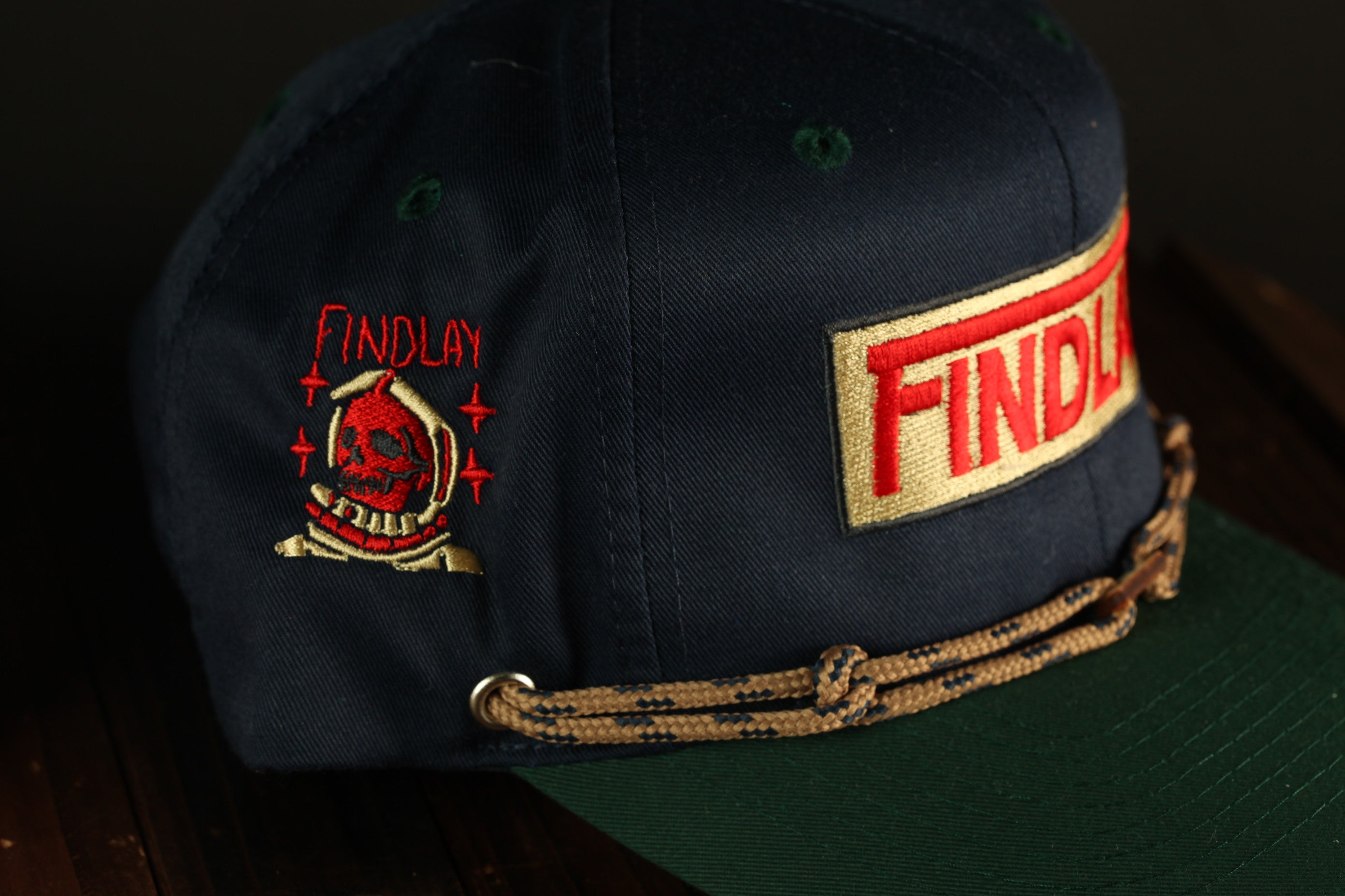 A dark blue baseball cap with a red and gold embroidered logo on the side and a brown braided strap.