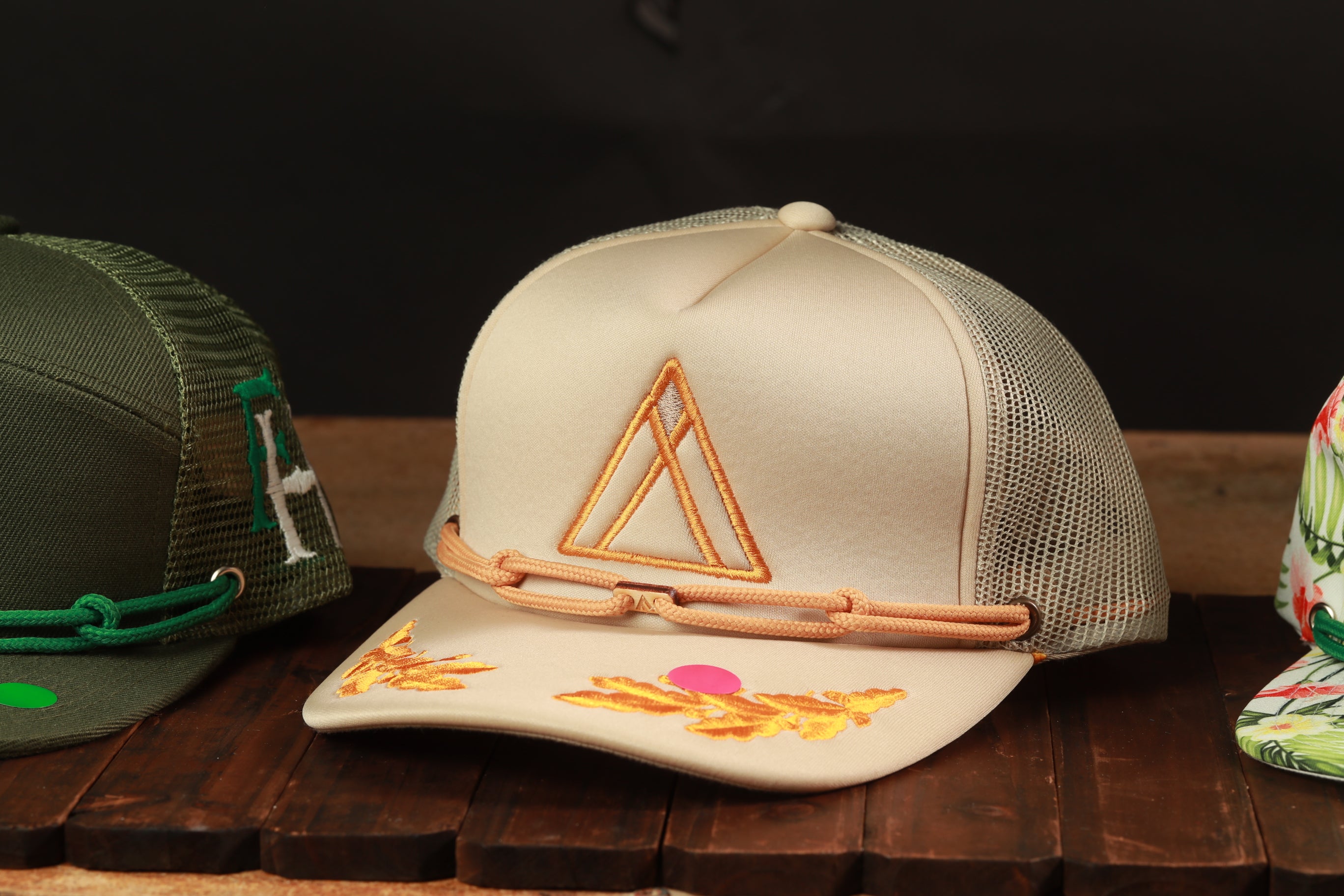 A beige baseball cap with a triangular logo and a chain strap is displayed on a wooden surface, with other hats visible in the background.