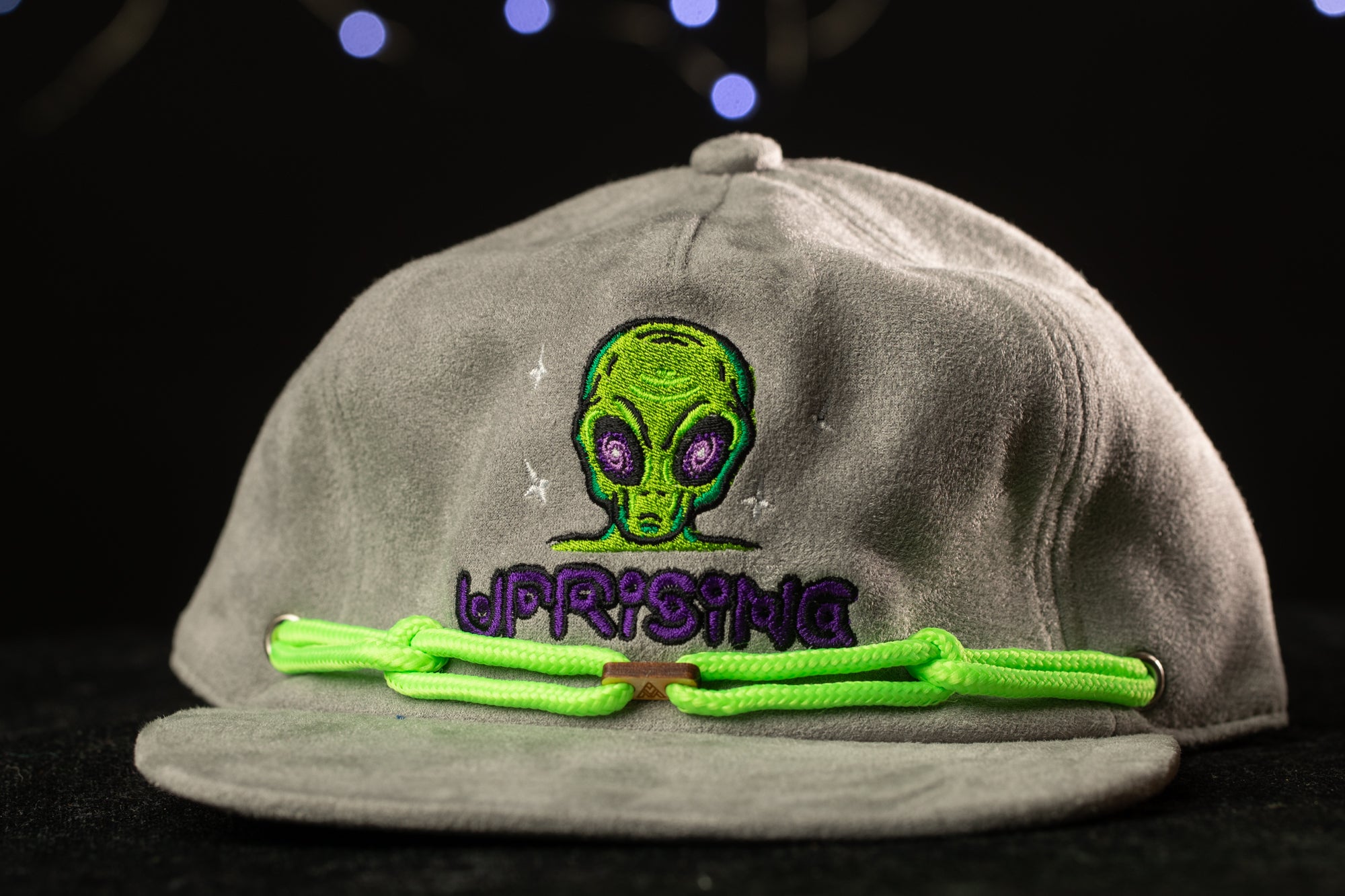 A gray baseball cap with a green alien head embroidered on it, and a green string or strap attached to the front.