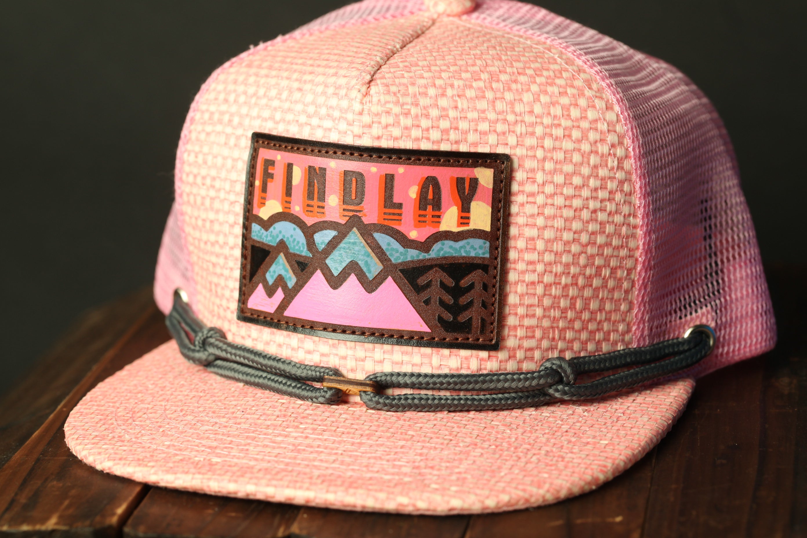 A pink baseball cap with a graphic patch on the front, featuring a mountain landscape design.