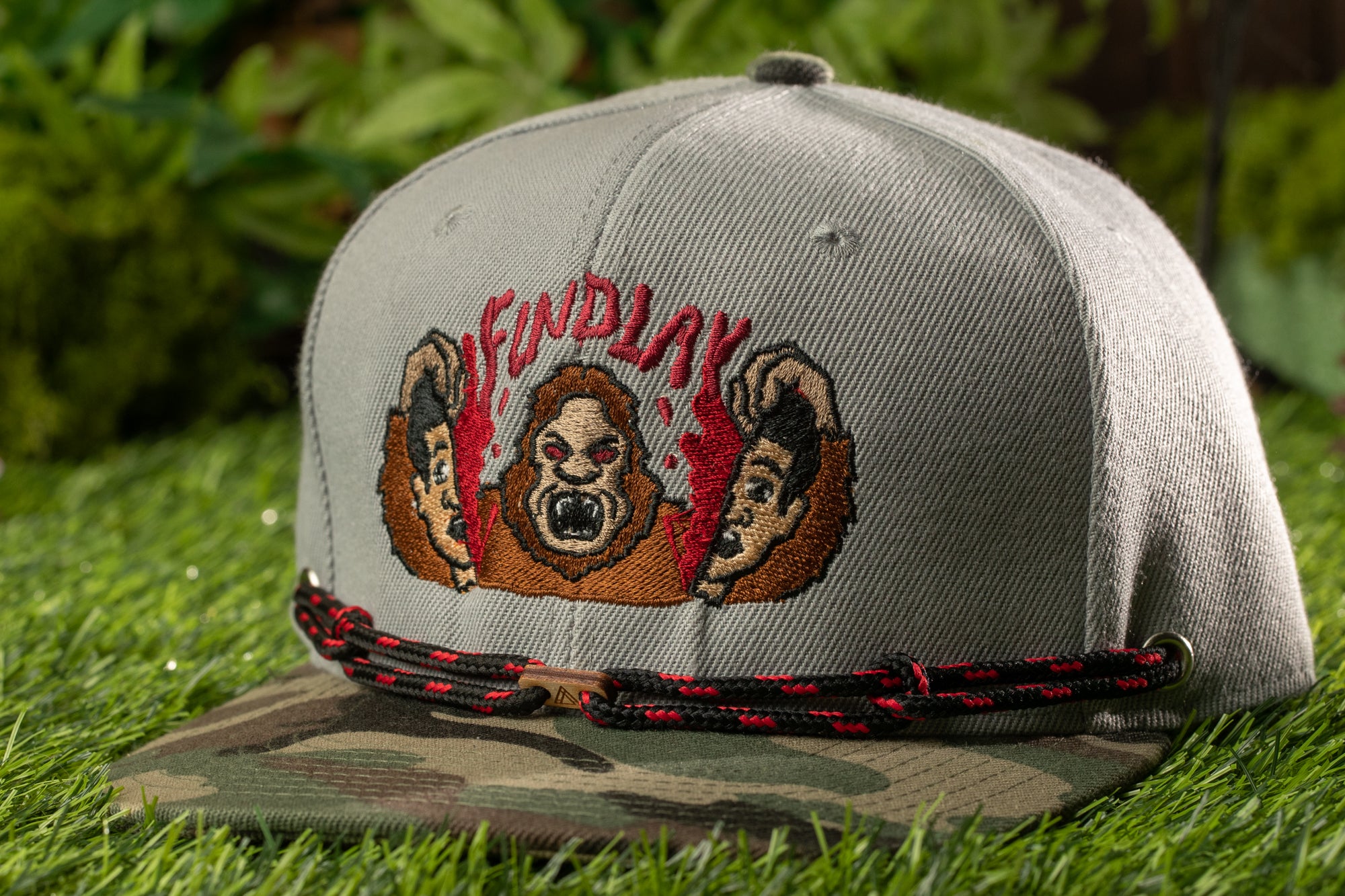 A gray baseball cap with a camouflage pattern and a graphic of a cartoon character on it, placed on a grassy surface.