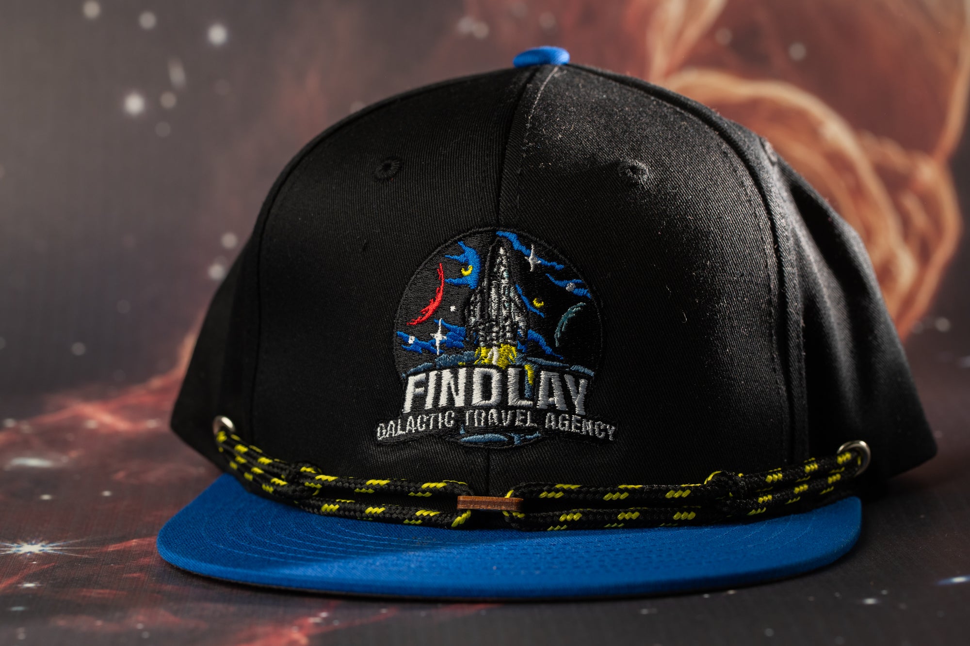 A black baseball cap with a blue brim and a logo on the front, against a space-themed background.