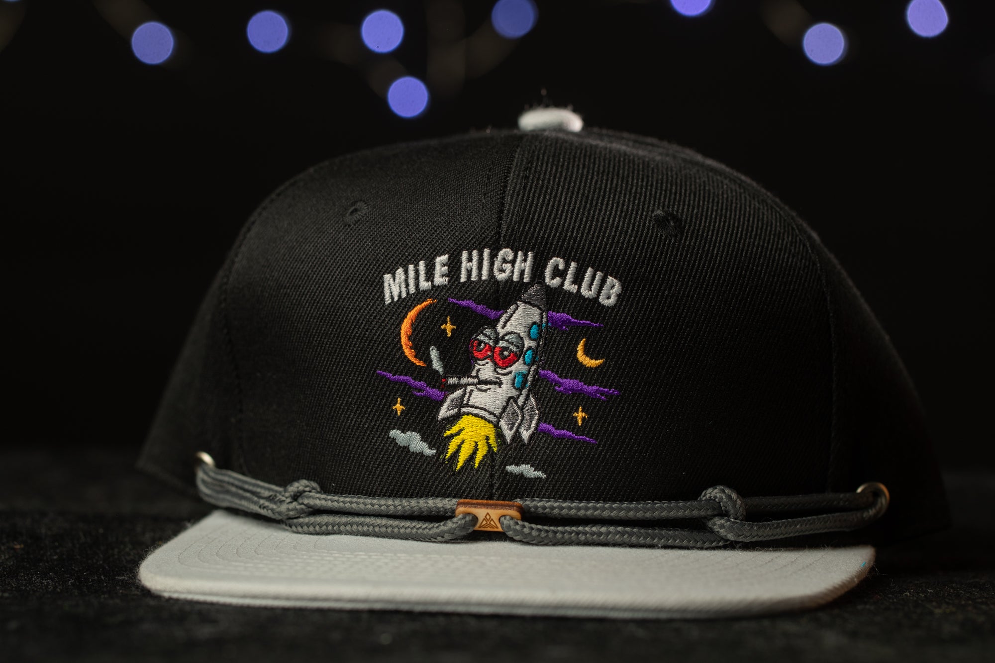 A black baseball cap with a colorful graphic and text on it, placed on a dark surface against a background with blue lights.