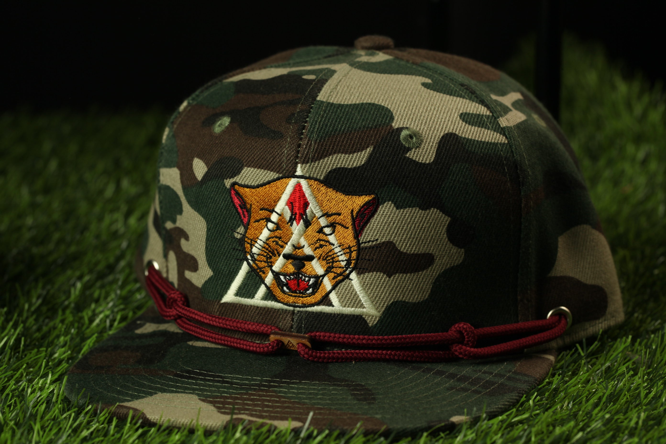 A camouflage baseball cap with a red chain strap and a cat-like logo on the front, placed on a grassy surface.