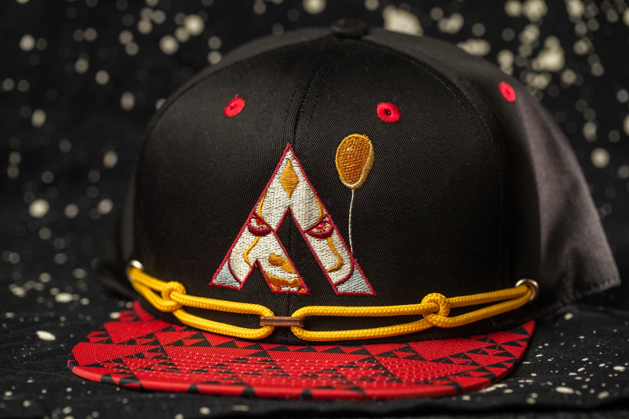 A black baseball cap with a red and gold geometric pattern, featuring a large "A" on the front and a yellow string around the brim.