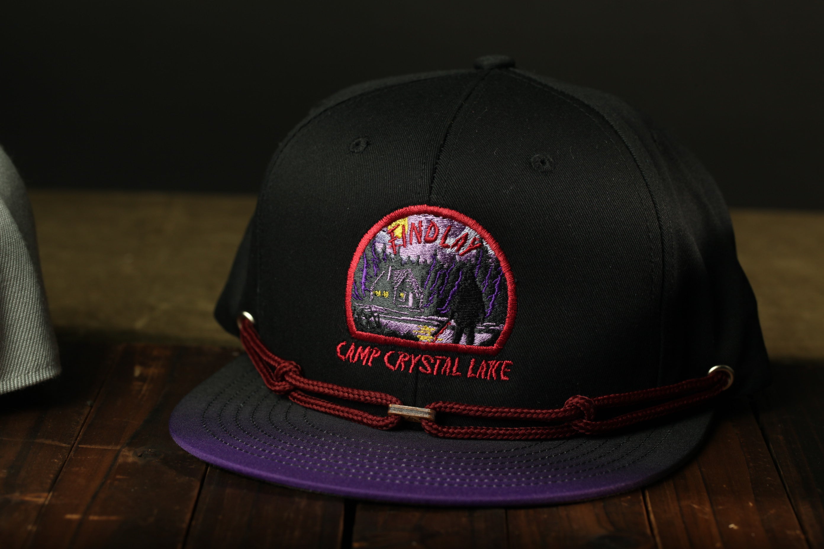 A black baseball cap with a red and purple embroidered logo, featuring the text "Camp Crystal Lake" on the front.