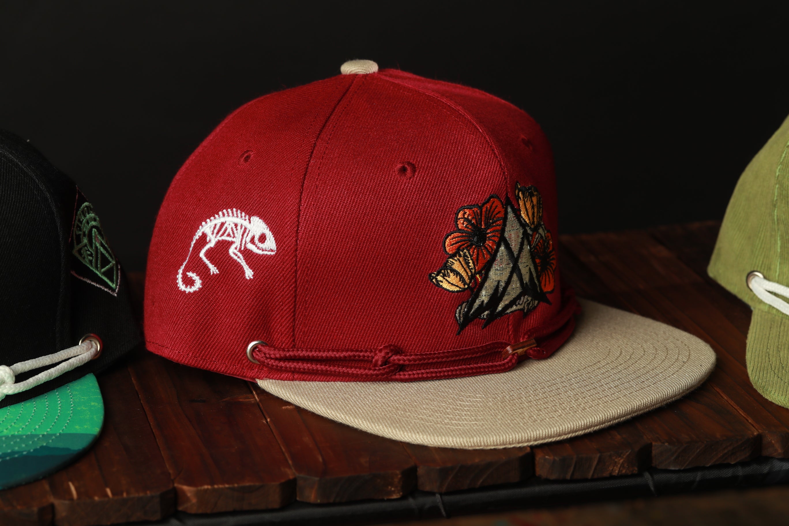 A red baseball cap with a white chameleon design and a floral embroidered patch is displayed on a wooden surface.