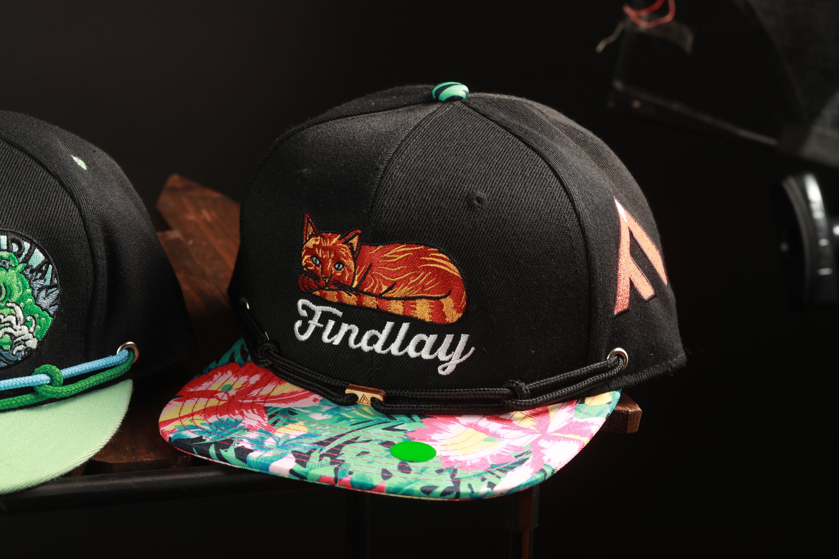 A close-up image of a black baseball cap with a colorful floral pattern and a cat embroidered on it.